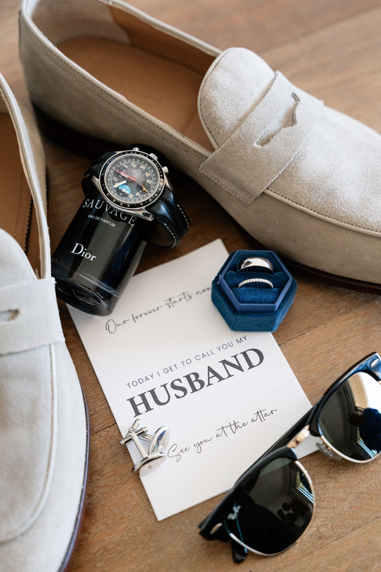 Elegant wedding accessories including white shoes, sunglasses, perfume bottle and invitation card arranged on wooden surface.