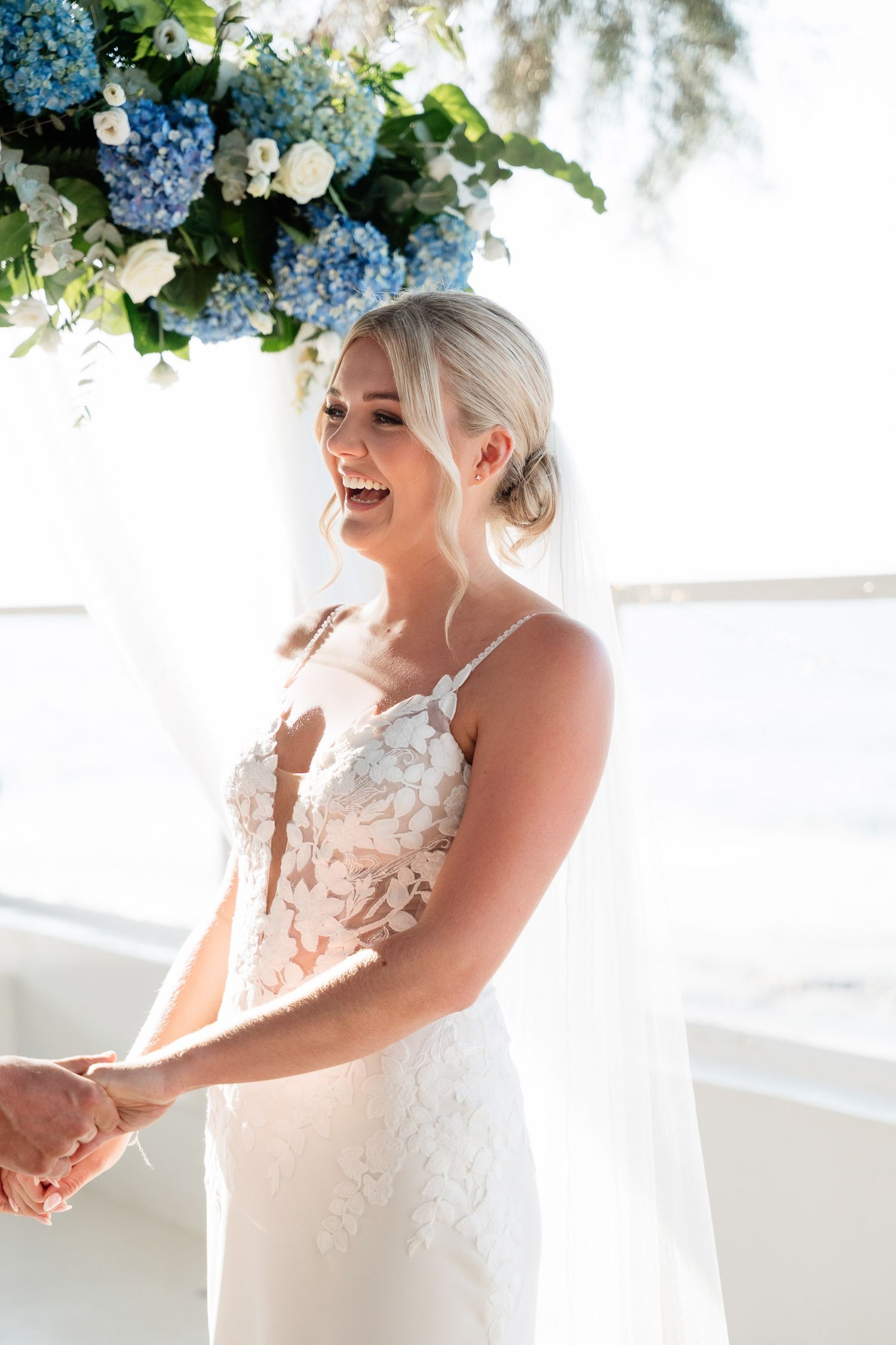 A bride wearing a delicate lace wedding dress laughs joyfully while holding blue hydrangea flowers on her special day.
