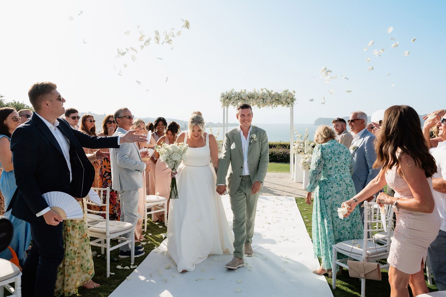 Newlyweds walk down outdoor aisle while wedding guests toss confetti during sunny beach ceremony.