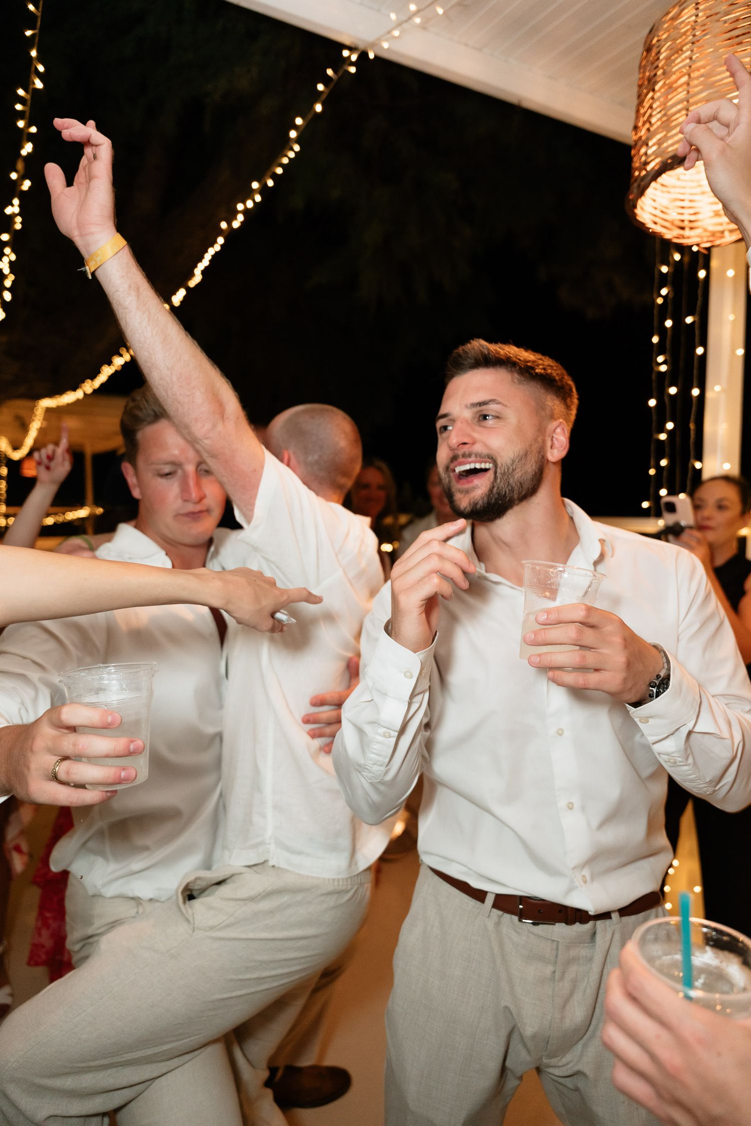 Party guests in white shirts dancing and celebrating with drinks under twinkling lights at a festive indoor event.