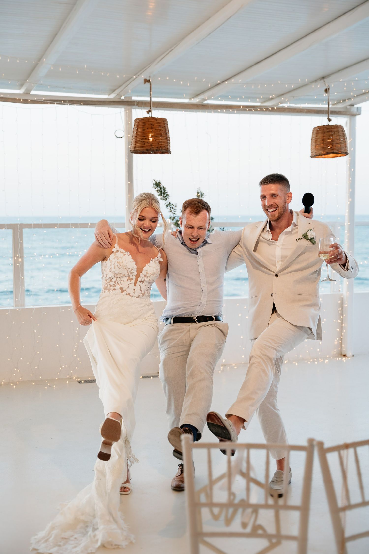 Three people dancing joyfully together at a beachside wedding reception under a white canopy with woven pendant lights.