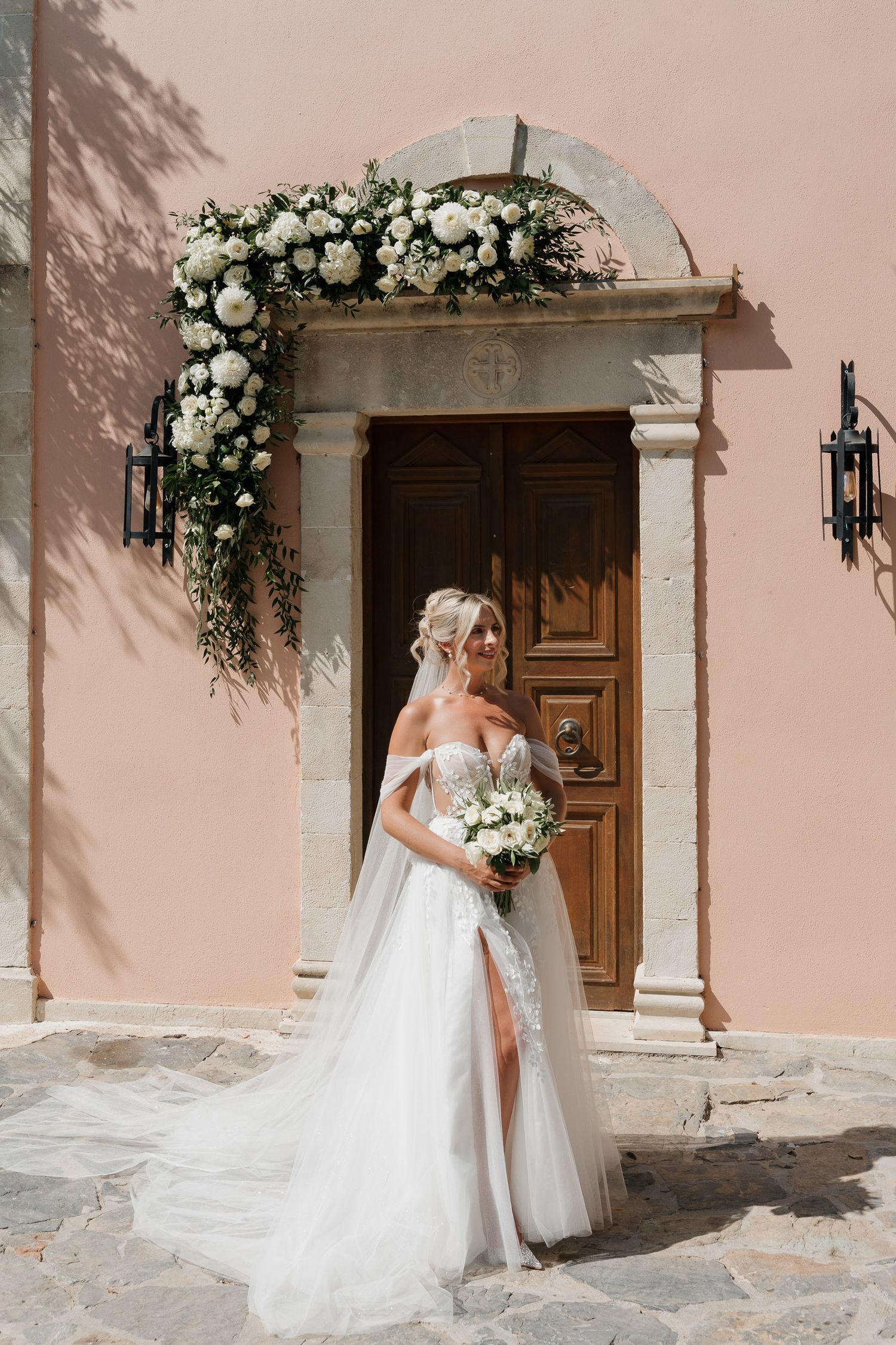 A bride in a flowing white wedding dress and veil poses in front of a rustic wooden door adorned with floral arrangements.