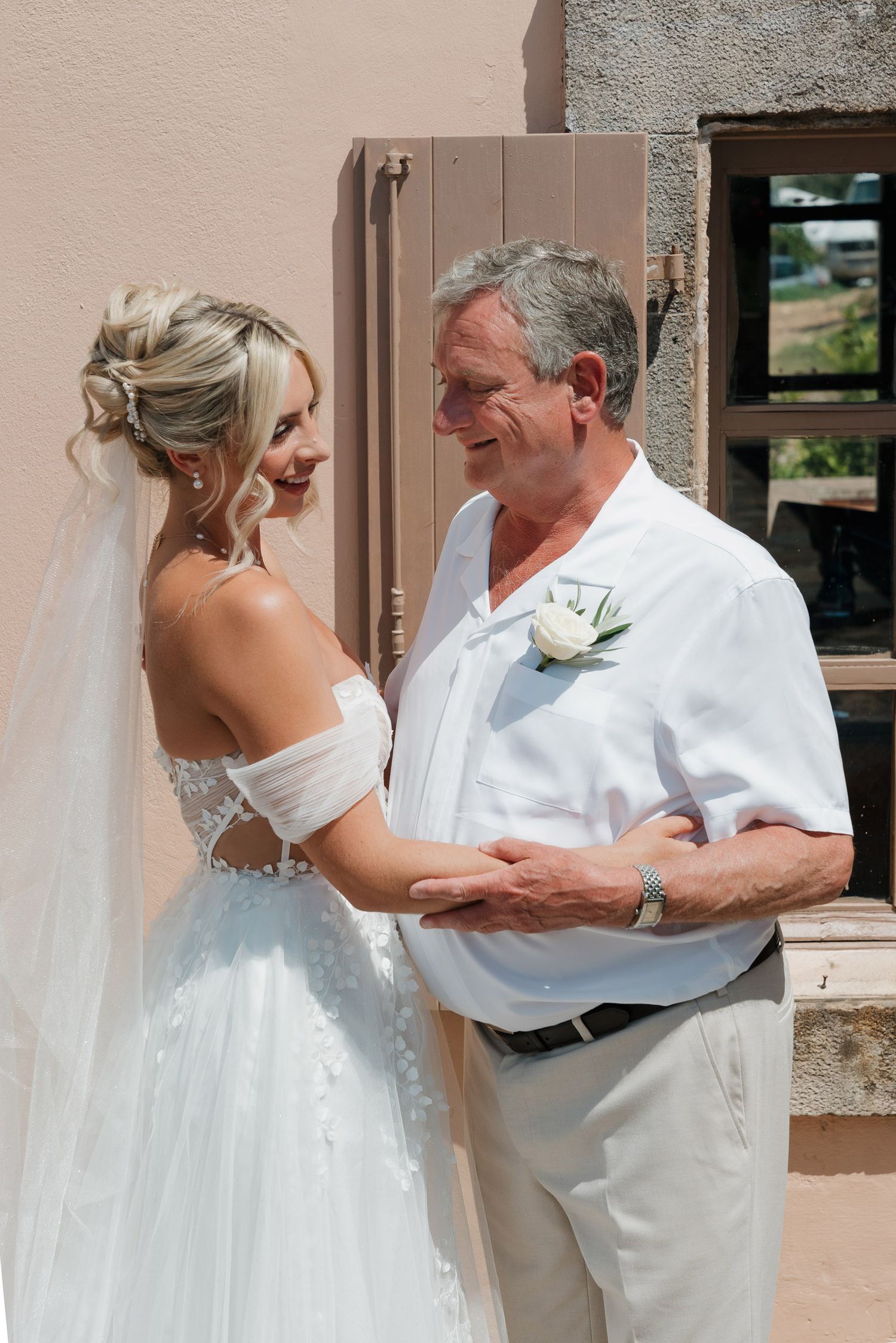 A tender moment between a bride in a white wedding dress and someone in white attire sharing an emotional embrace outdoors.