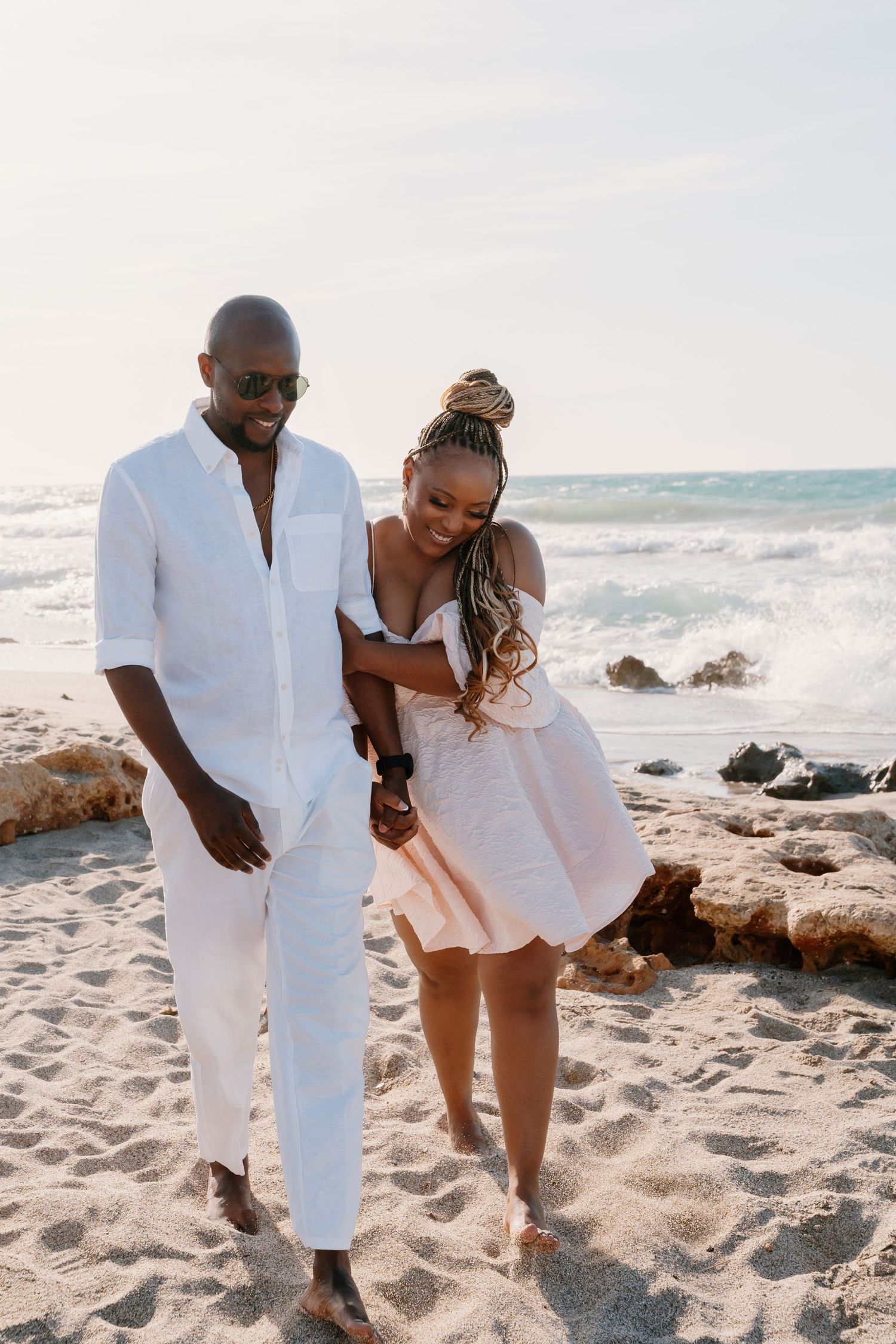 A couple in white outfits shares playful moments together on a rocky beach at sunset.