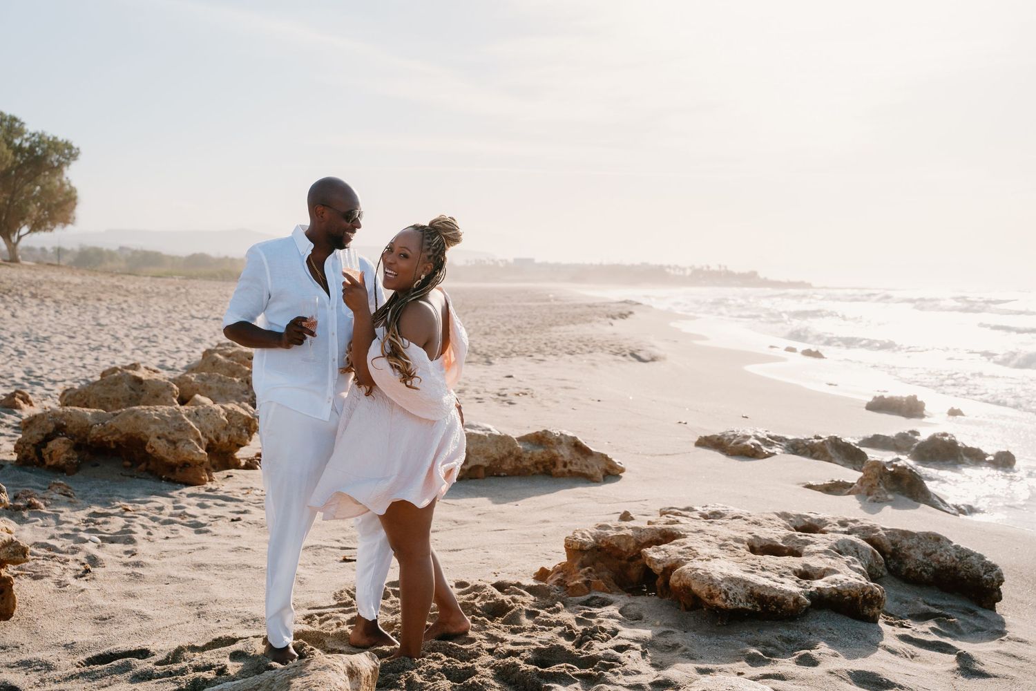 A couple in white outfits walks together along a rocky beach shoreline at sunset.