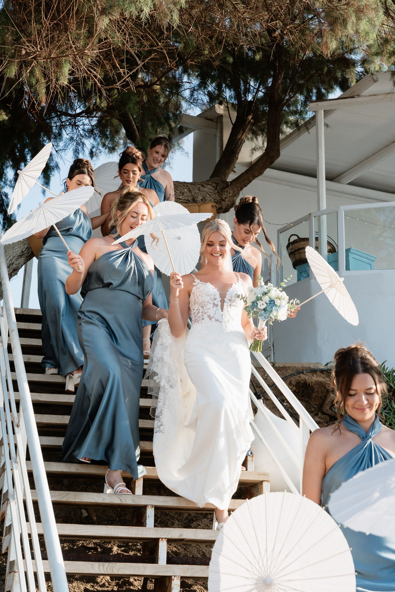 Wedding party in blue and white attire poses playfully with parasols on outdoor stairs during sunny celebration.
