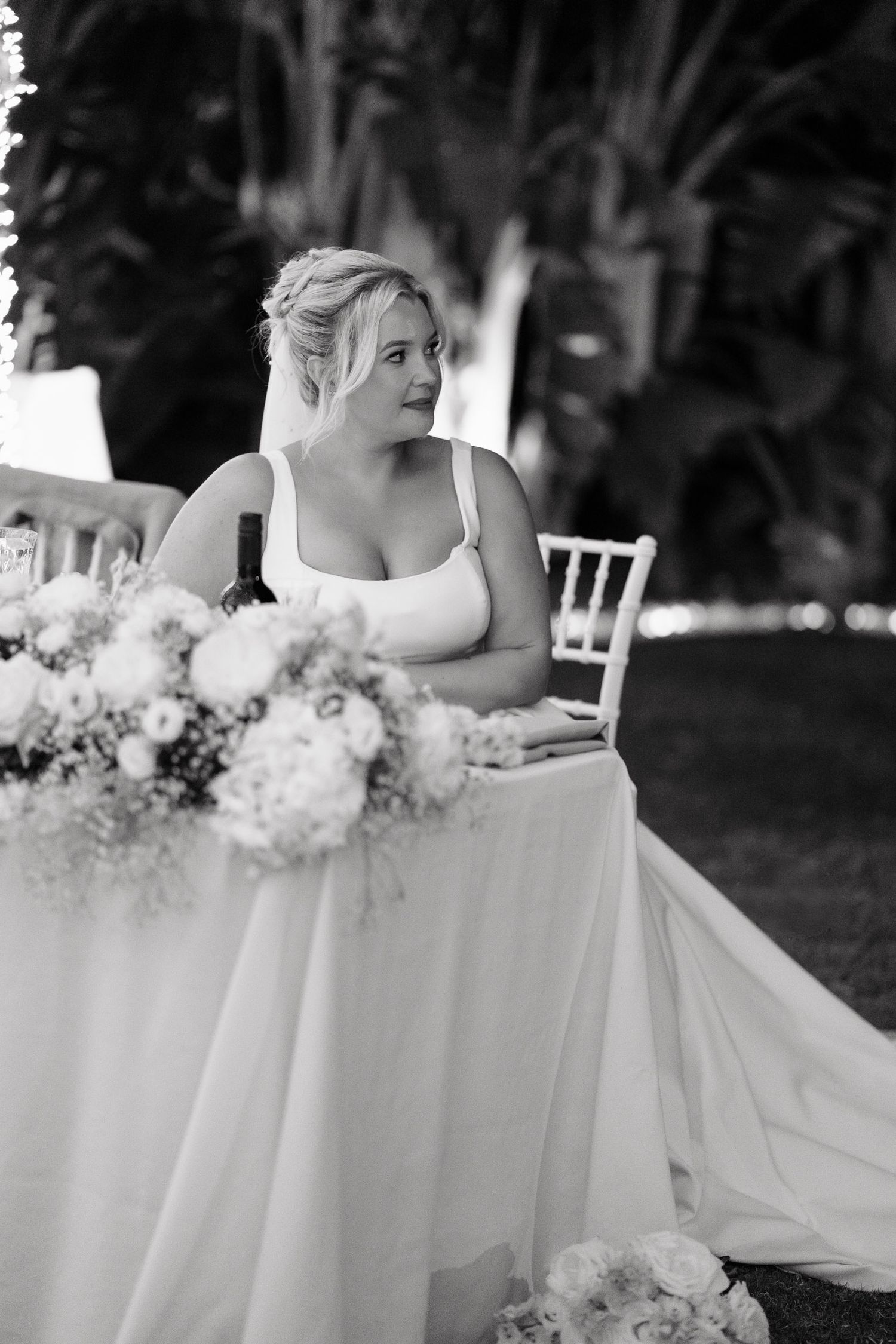 Black and white wedding photo showing a bride seated at a decorated table with white floral arrangements and flowing tablecloth.