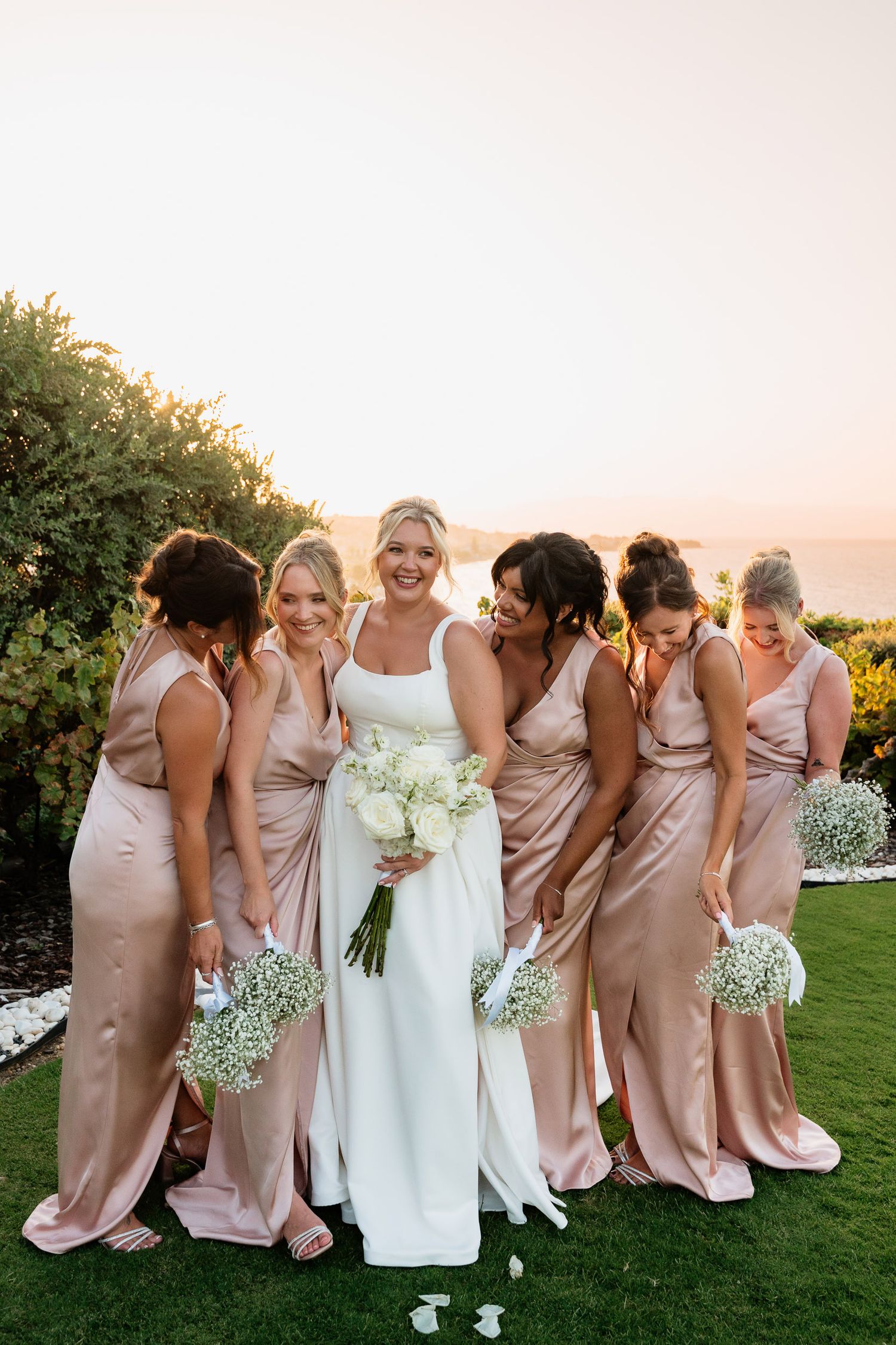 A bridal party in blush pink dresses poses together on a grassy lawn at sunset for candid wedding photos.