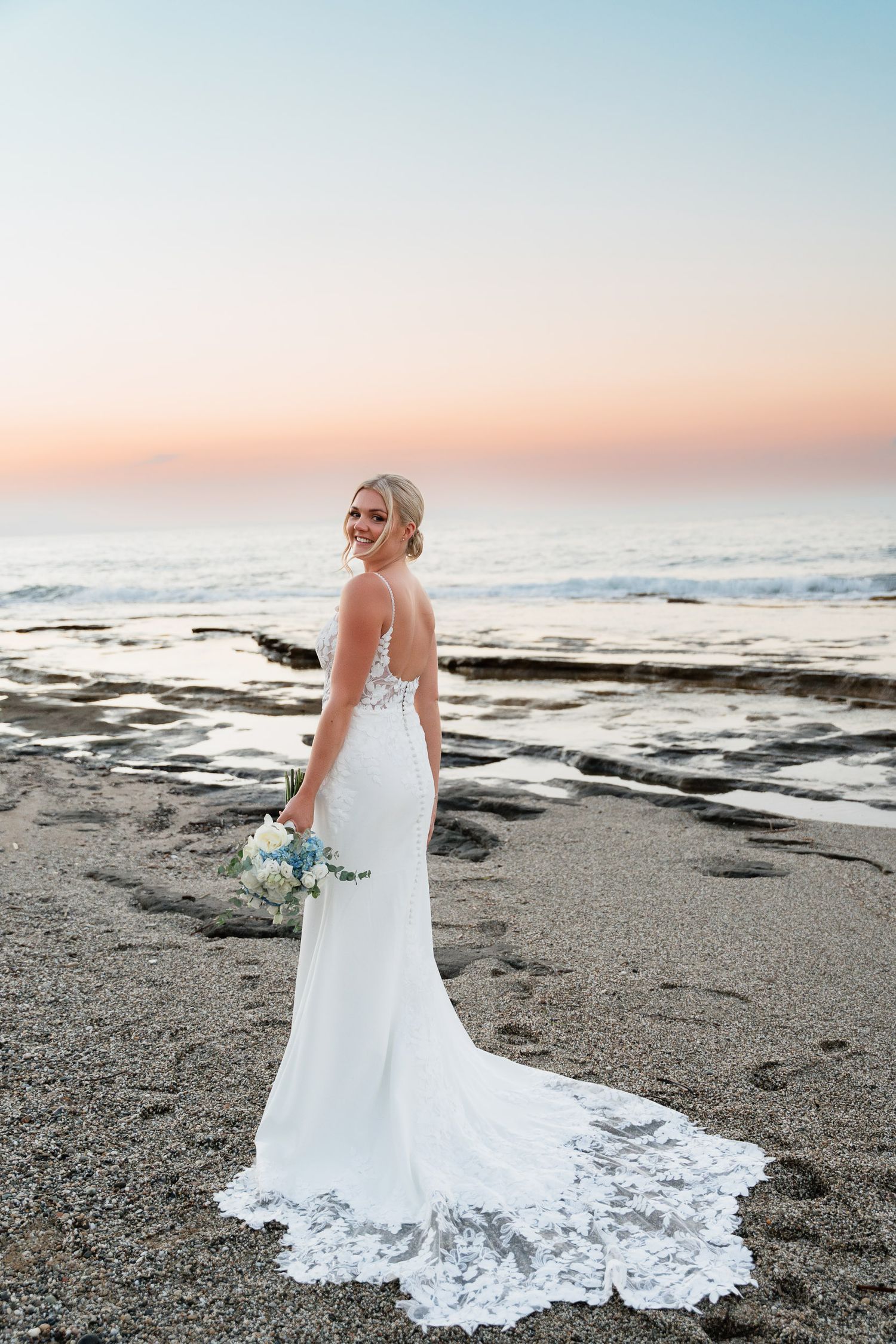A beautiful beach wedding portrait shows a bride in a flowing white lace gown against the ocean at sunset.