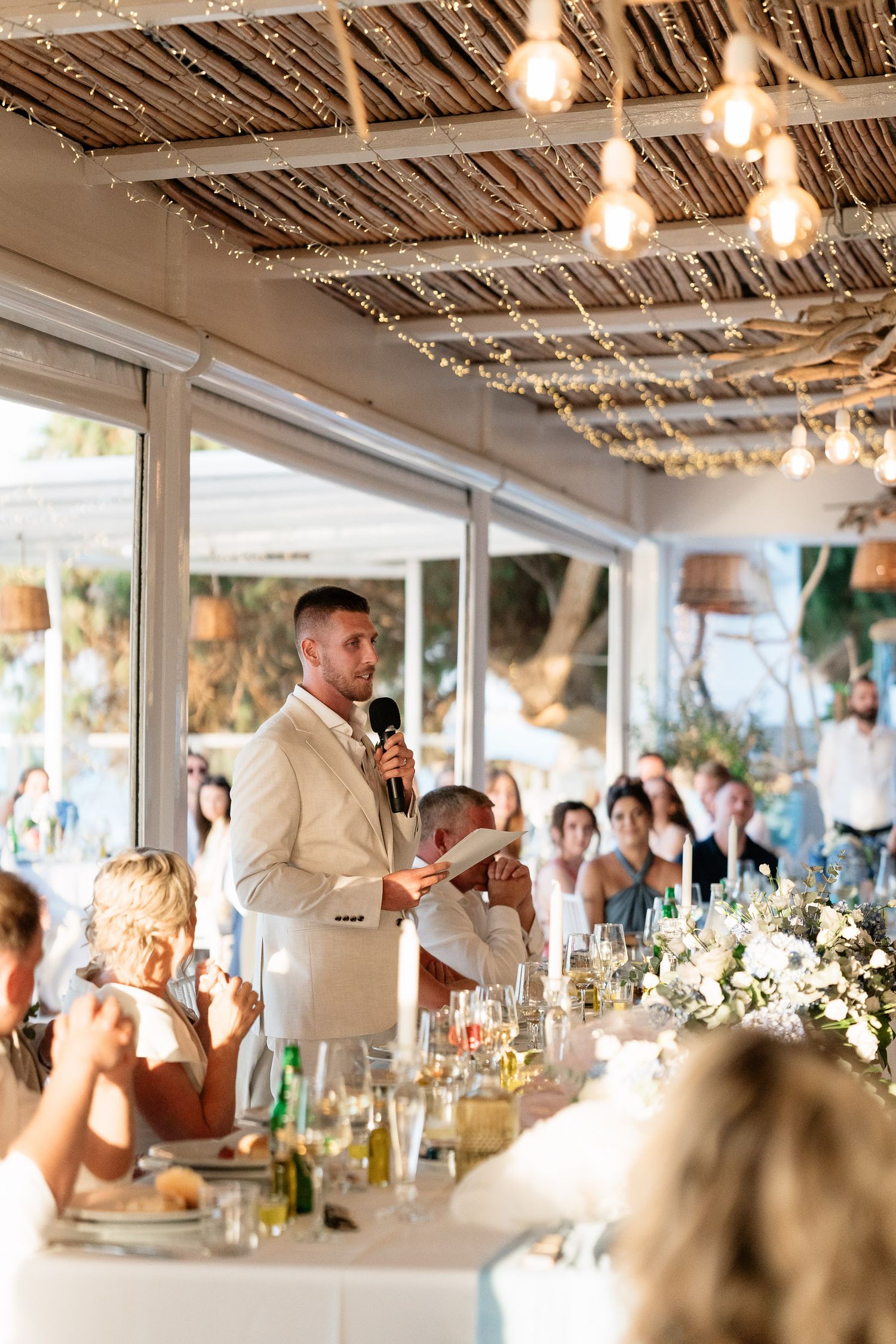 Wedding guests gather in an elegant reception venue with twinkling lights and white floral decorations on tables.
