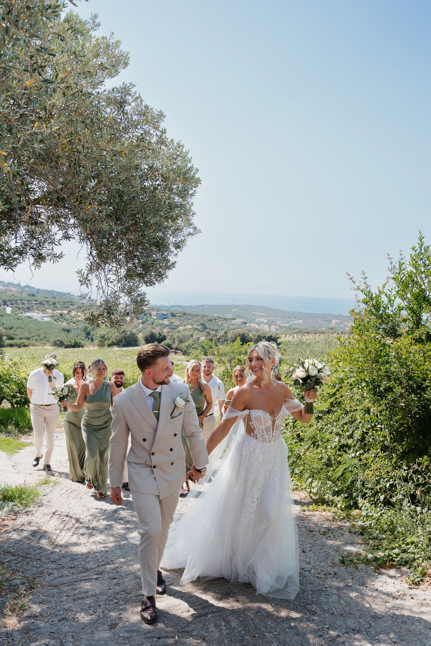 Wedding guests gather under an olive tree on a scenic hillside overlooking a Mediterranean landscape during a summer celebration.