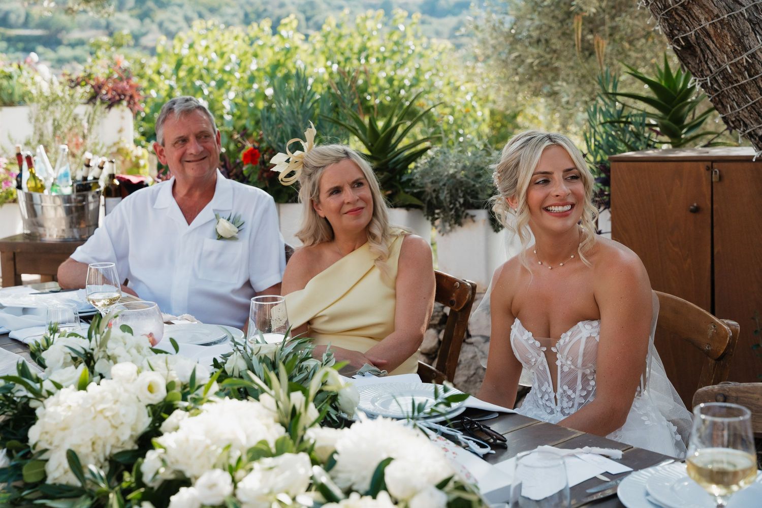 Wedding guests seated at outdoor reception tables share joyful moments amid white floral centerpieces and garden scenery.
