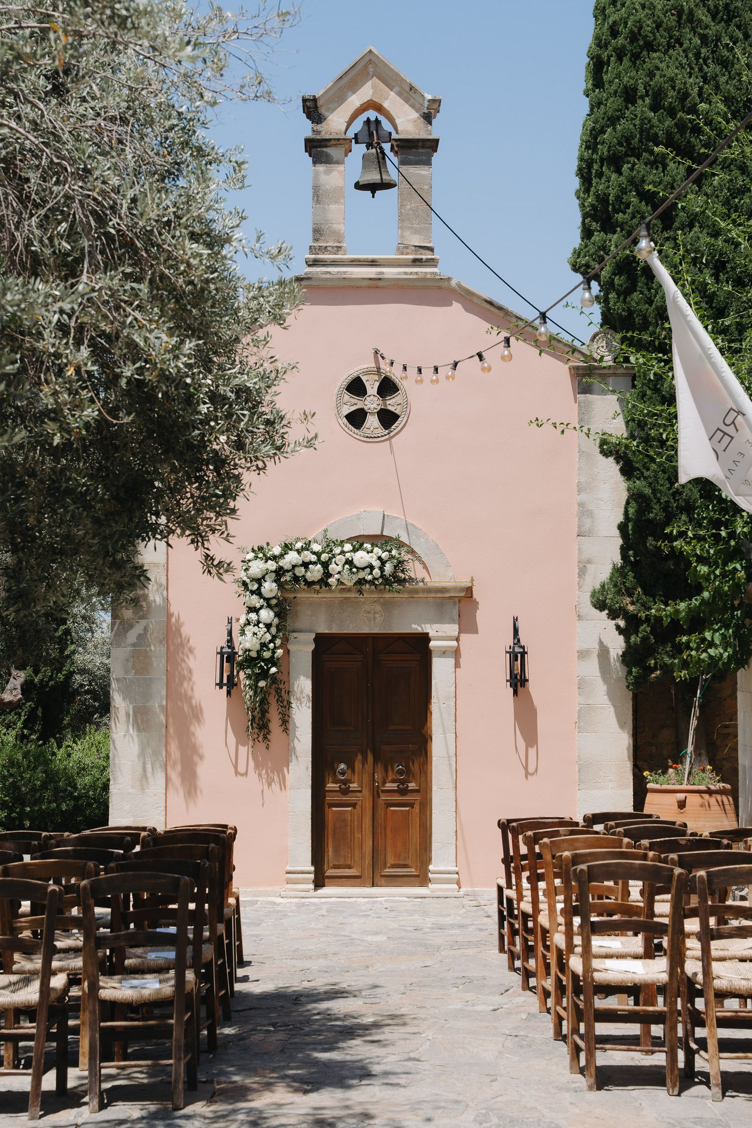 Pink Mediterranean chapel with wooden doors, floral arch, and rows of wooden chairs set up for an outdoor wedding ceremony.