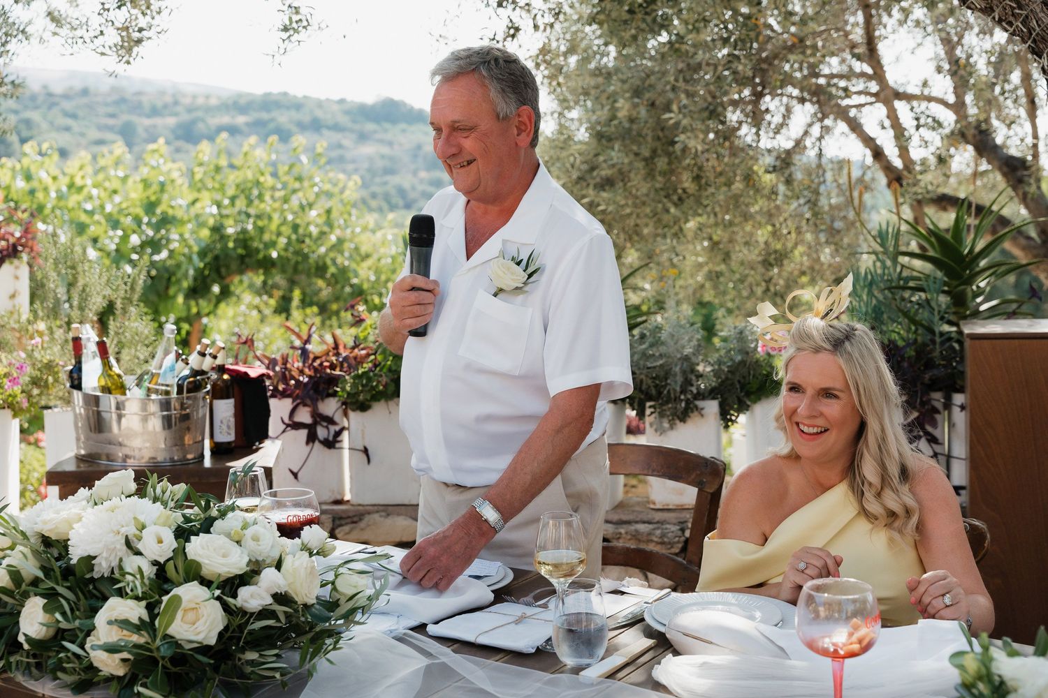 A wedding reception speech being given outdoors at an elegant table setting with white floral arrangements and olive trees.