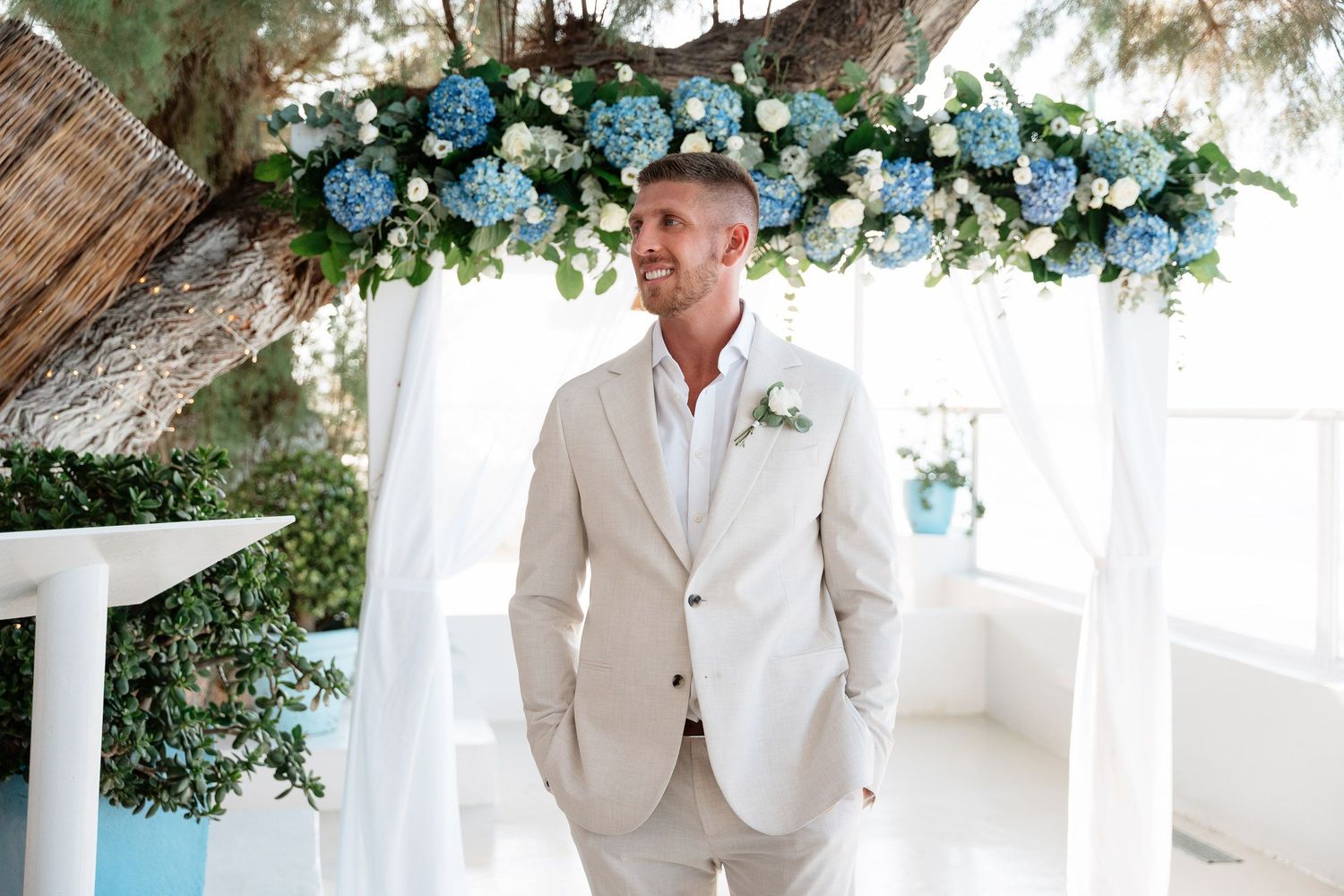 A groom in a light beige suit stands under an elegant floral arch with blue hydrangeas at a beach wedding ceremony.