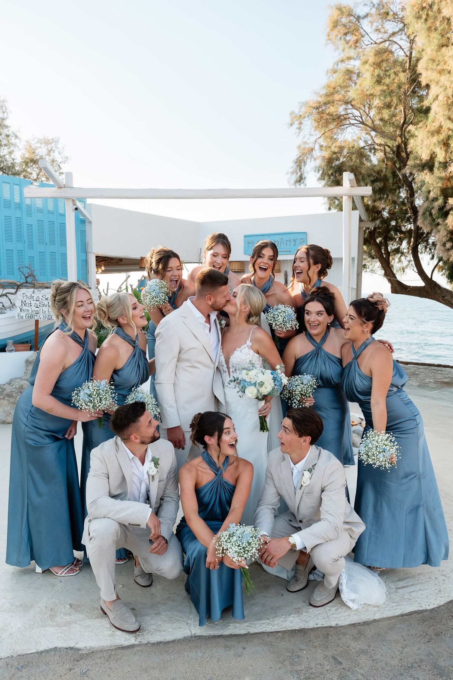 Wedding party in light blue bridesmaid dresses and light colored suits poses together on a sunny beach.