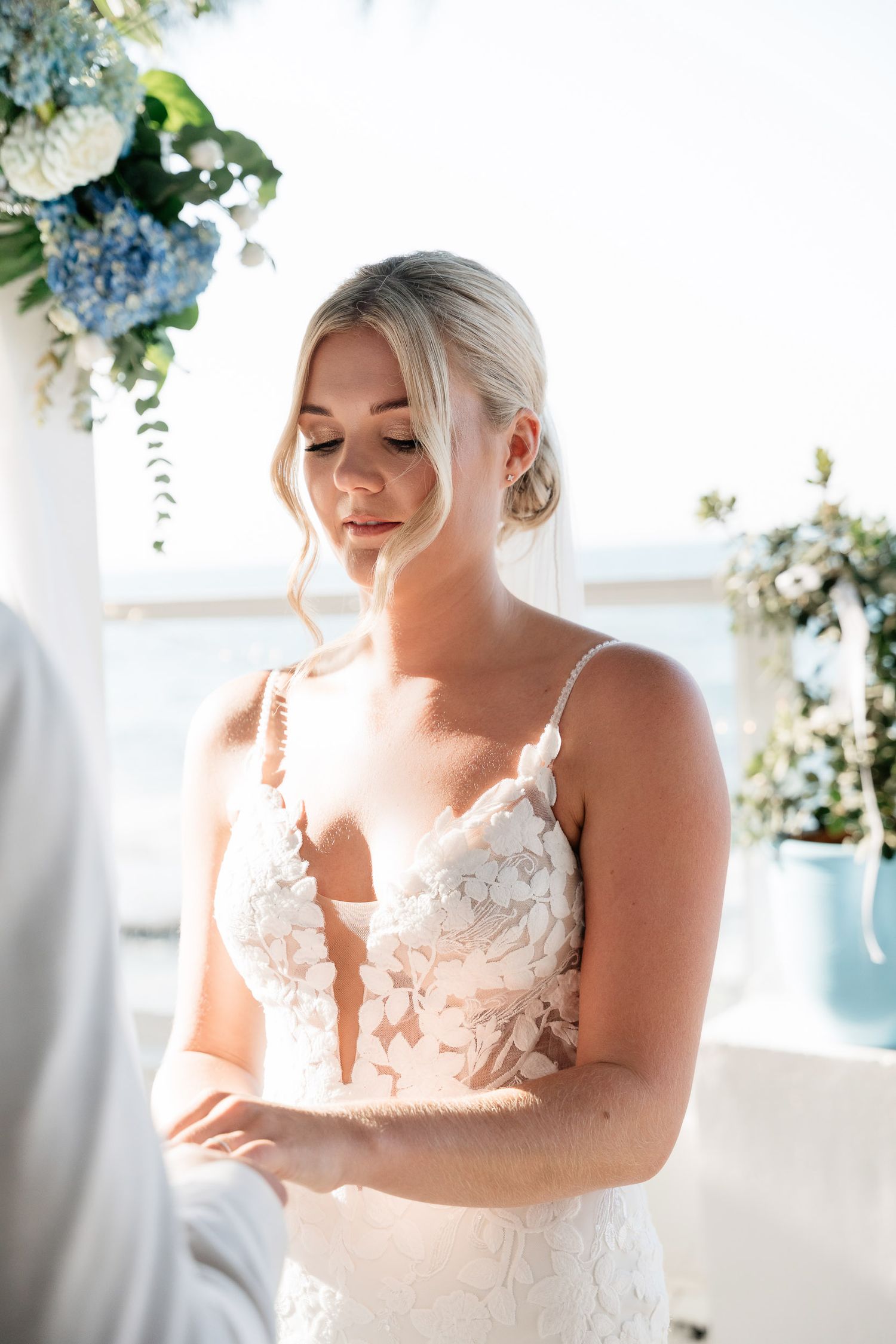 A bride in a delicate lace wedding gown stands during a ceremony with floral arrangements in soft blues and whites.