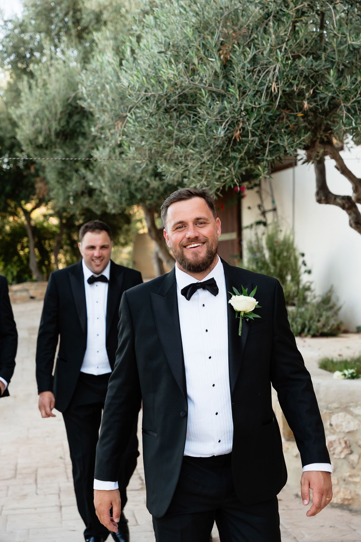 A group in formal black tuxedos and bow ties walk and laugh together on a stone path surrounded by olive trees.