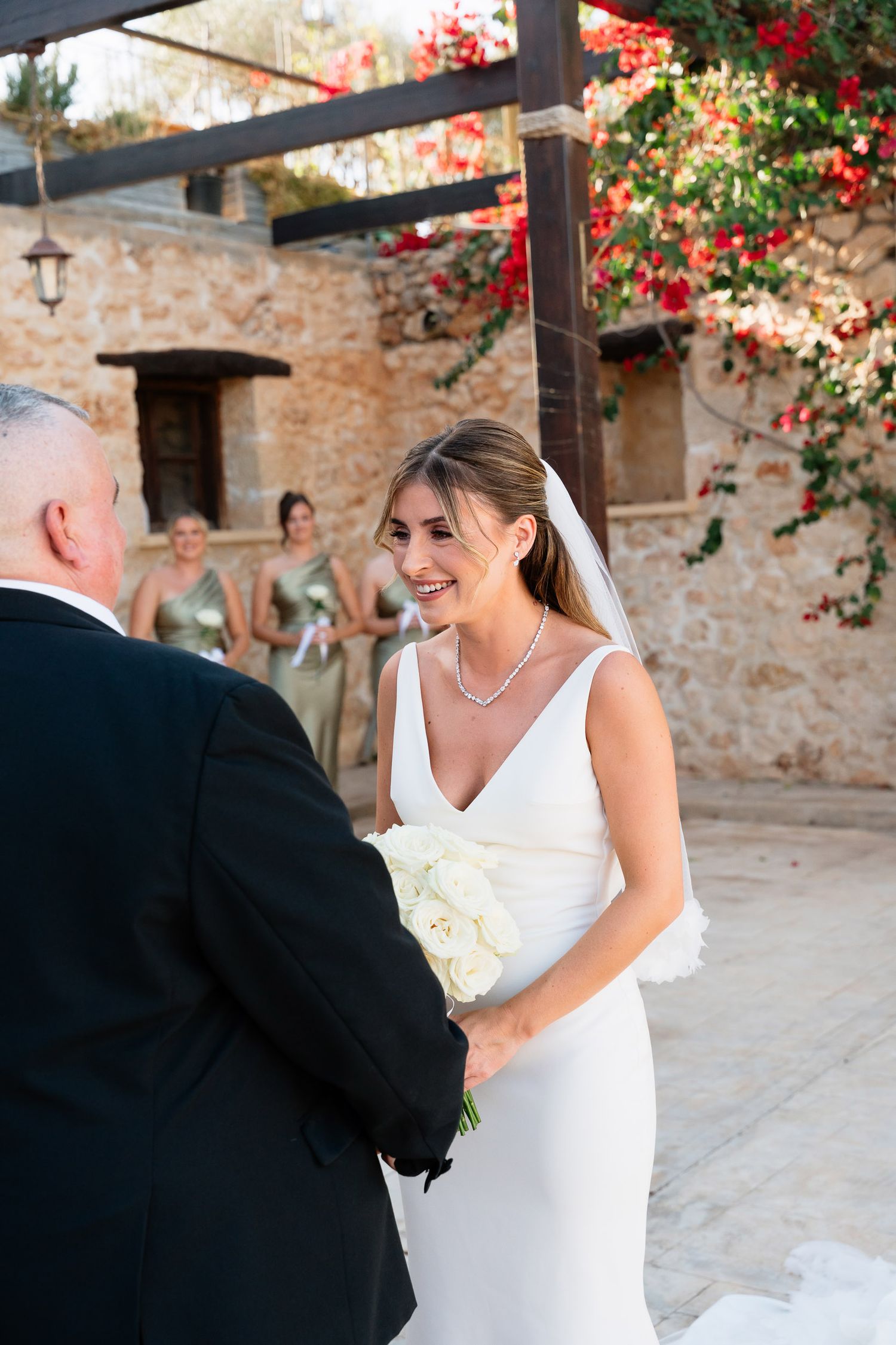 A bride in a white wedding dress shares a joyful moment with guests in a rustic stone courtyard with red flowers above.