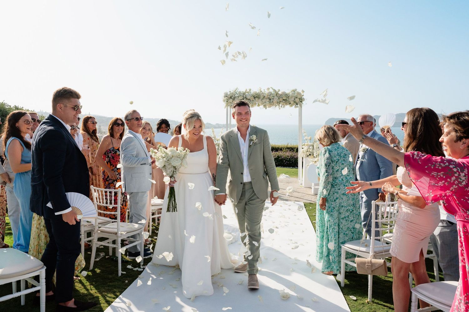 Wedding ceremony on a sunny beach with guests tossing flower petals as newlyweds walk down white aisle runner.