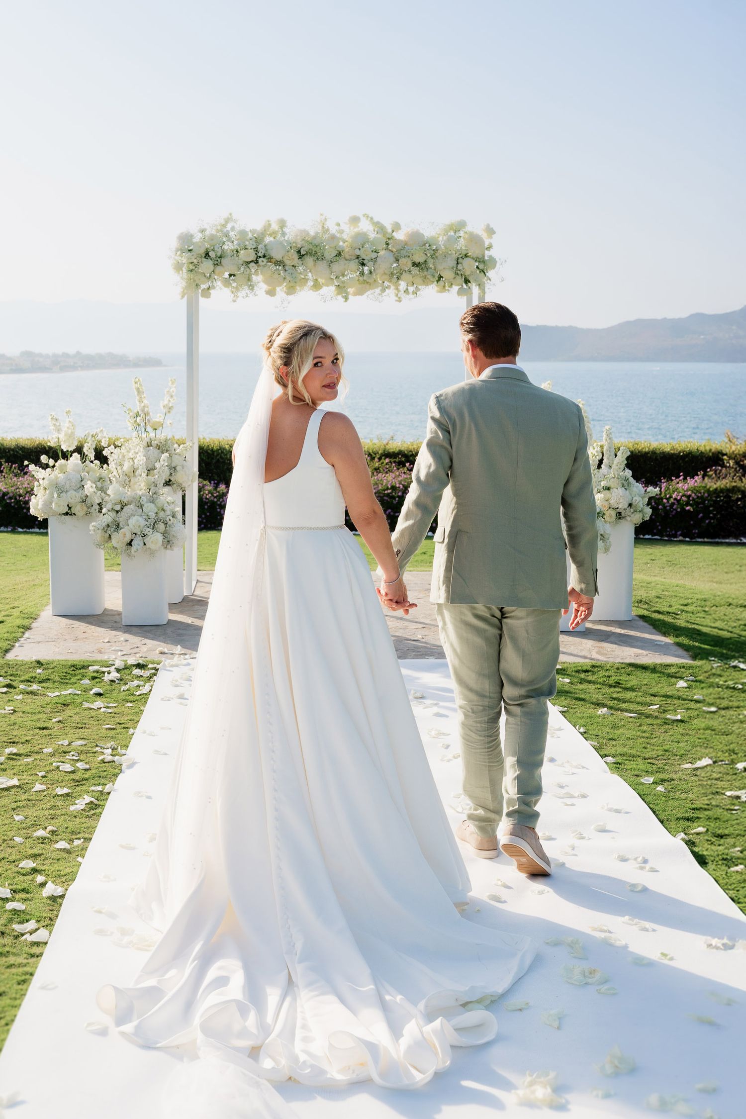 A couple in wedding attire stands at a scenic oceanfront ceremony site with white floral decorations and mountain views.