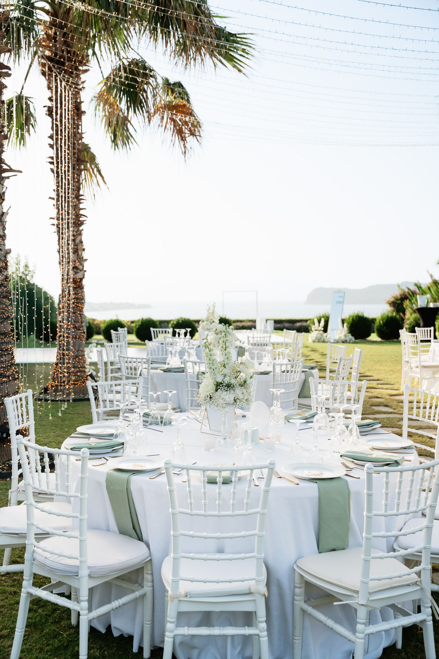 Elegant outdoor wedding reception setup with white Chiavari chairs and round tables under palm trees by the ocean.
