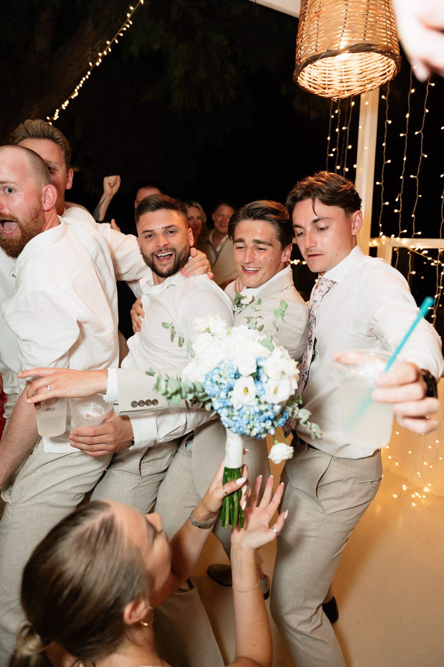 Groomsmen in light colored suits celebrate at a wedding reception with a blue and white bouquet under string lights.