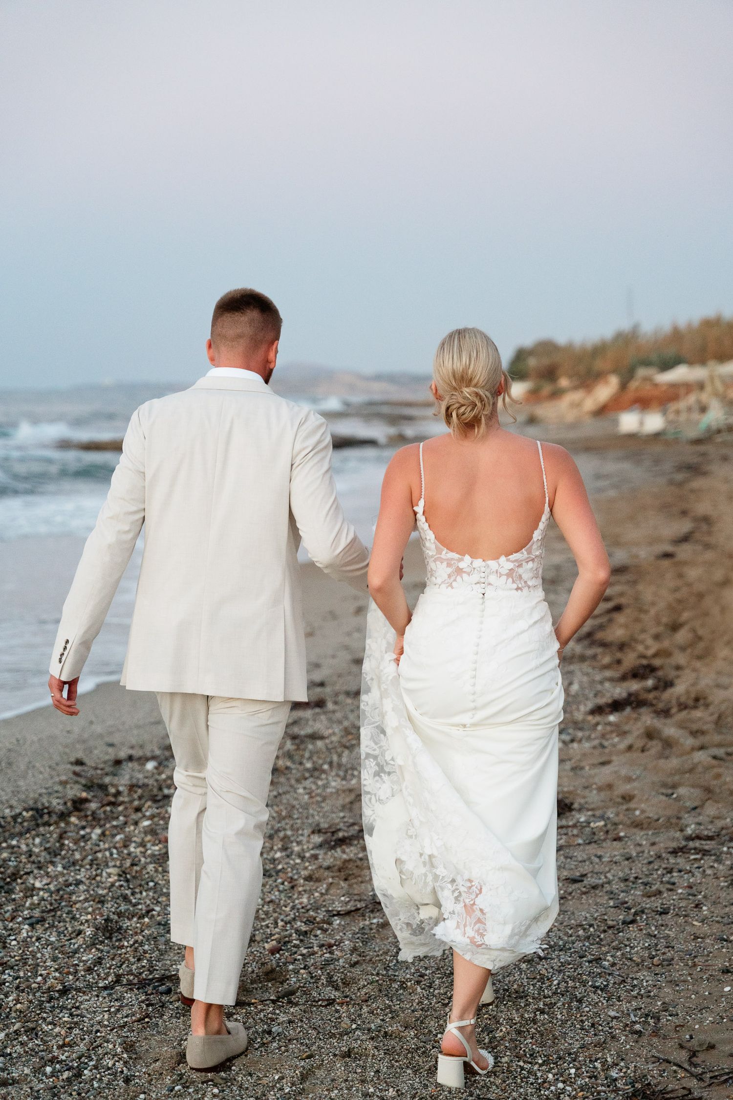 A couple in white wedding attire walks together along a rocky beach at sunset with waves crashing in the background.