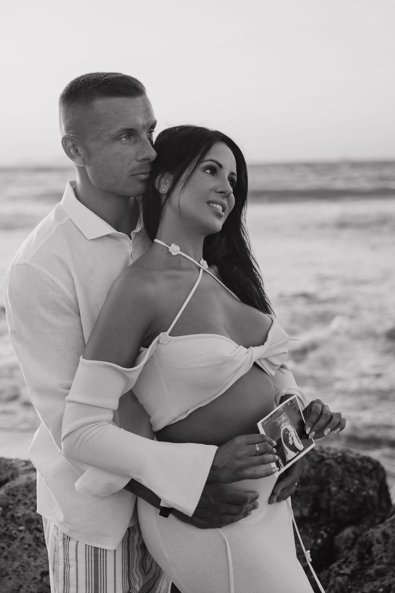 A romantic black and white photo series of a couple embracing on a rocky beach at sunset.