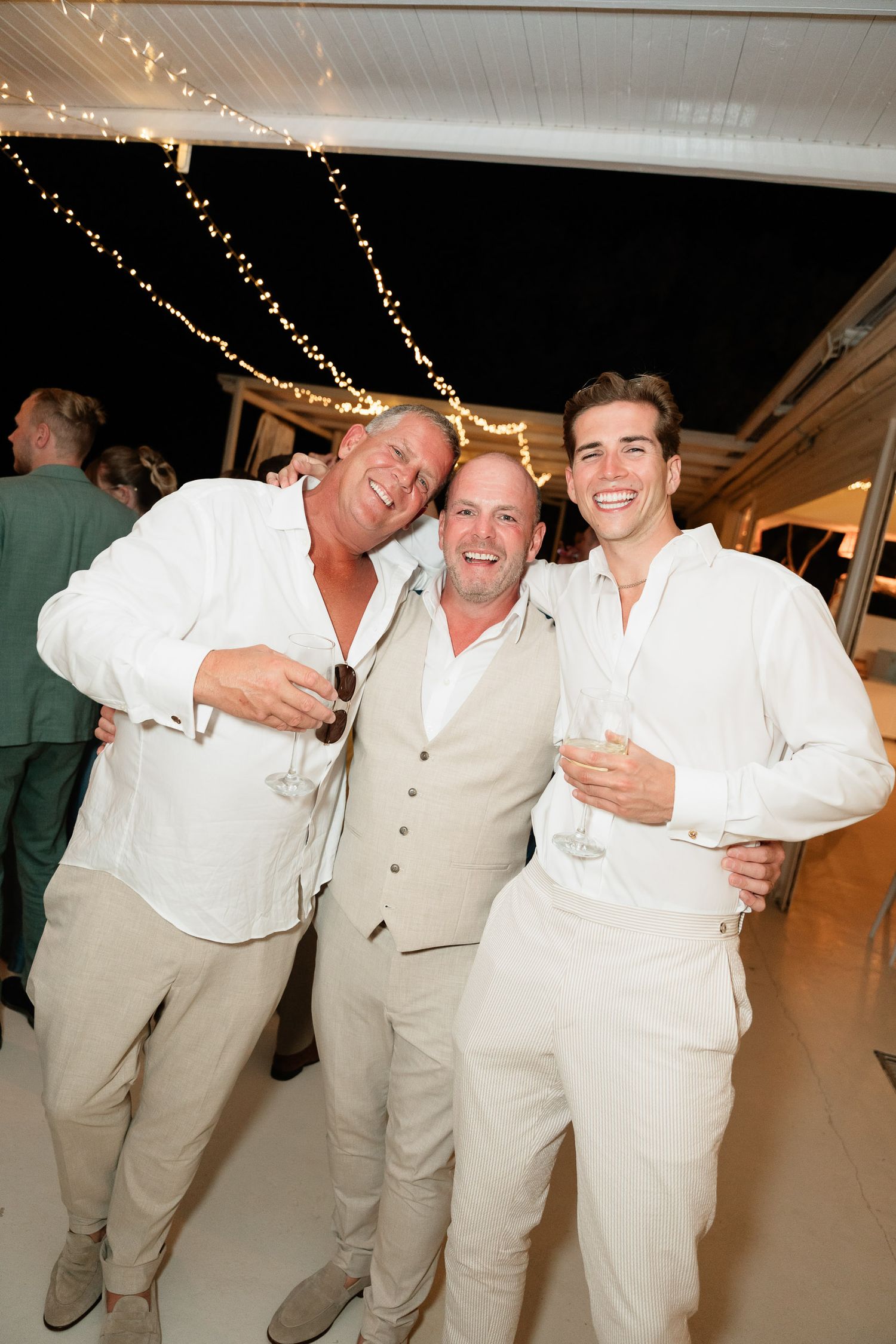 Three people in light colored attire smile and pose together at an indoor celebration with string lights overhead.
