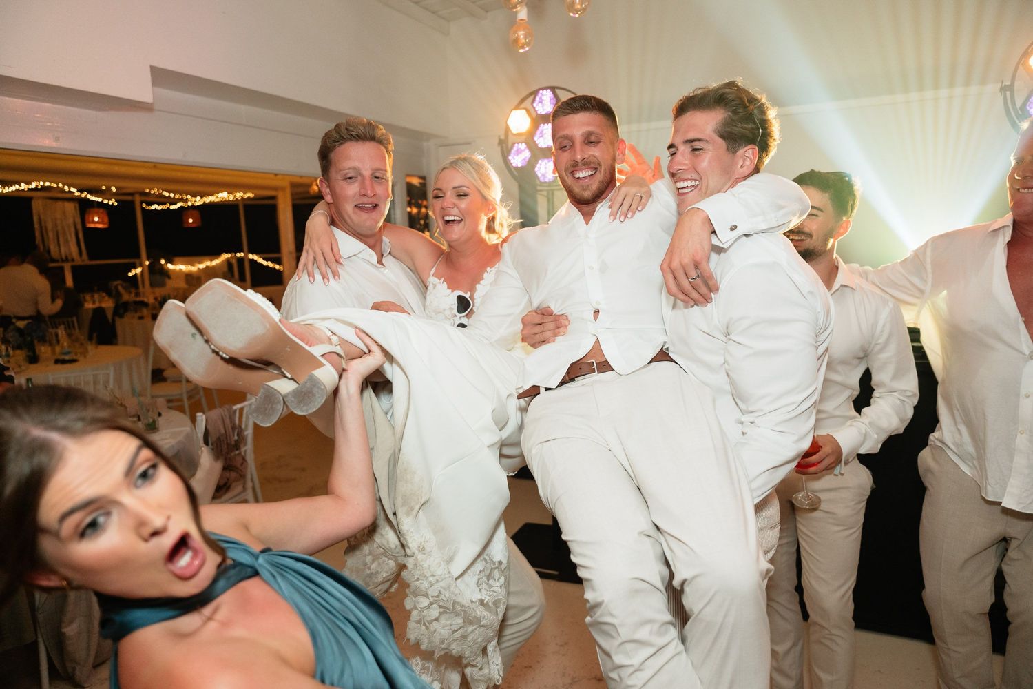 Wedding party in white attire dancing and celebrating joyfully on the dance floor at a reception.