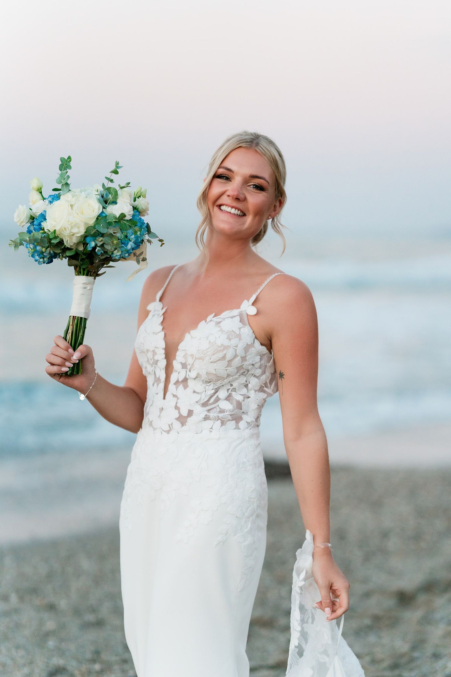A bride in a white lace wedding dress poses joyfully on a beach with a blue and green floral bouquet.