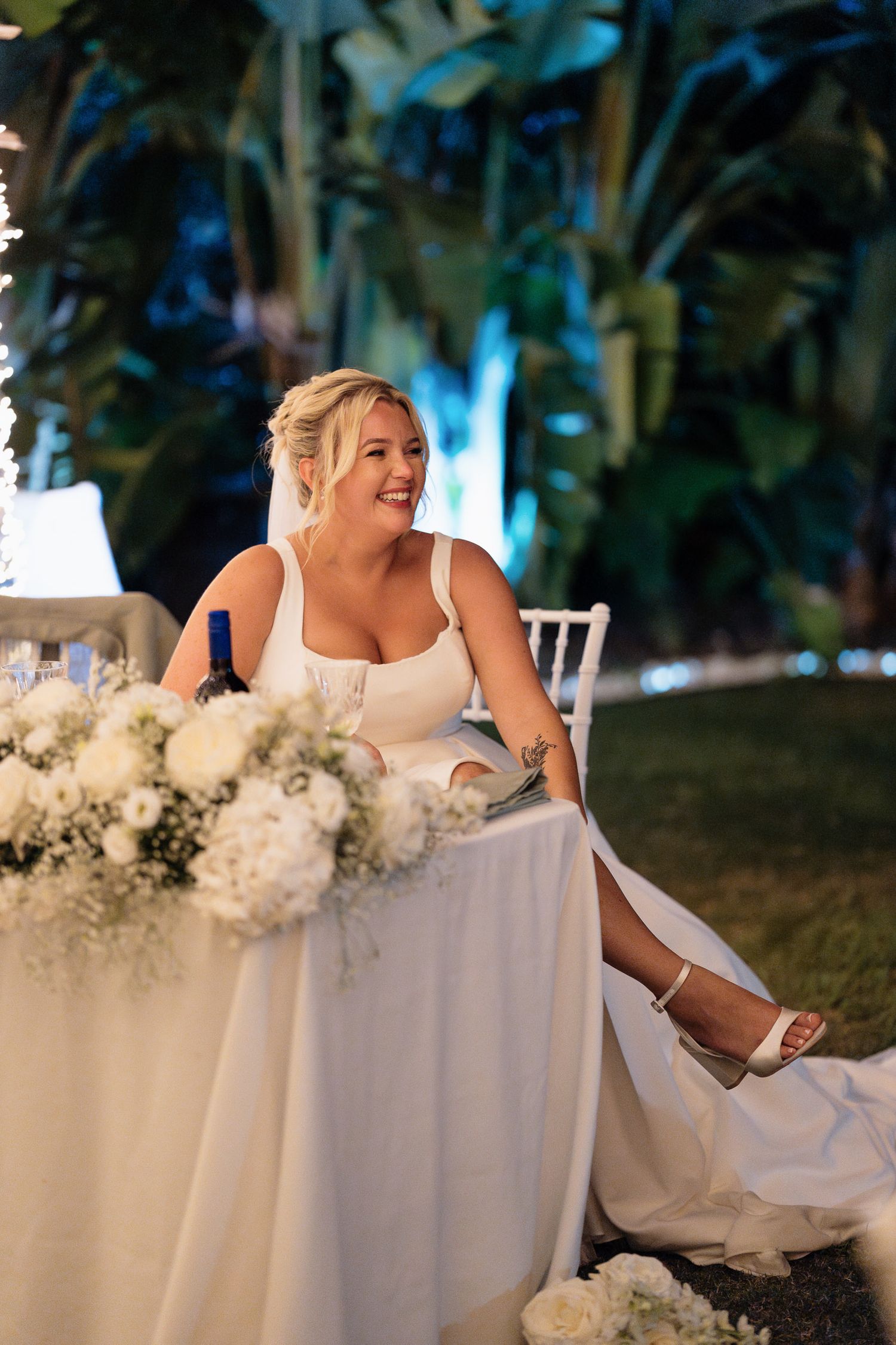 A bride in a white wedding dress sits at an elegantly decorated table with floral arrangements during an evening reception.