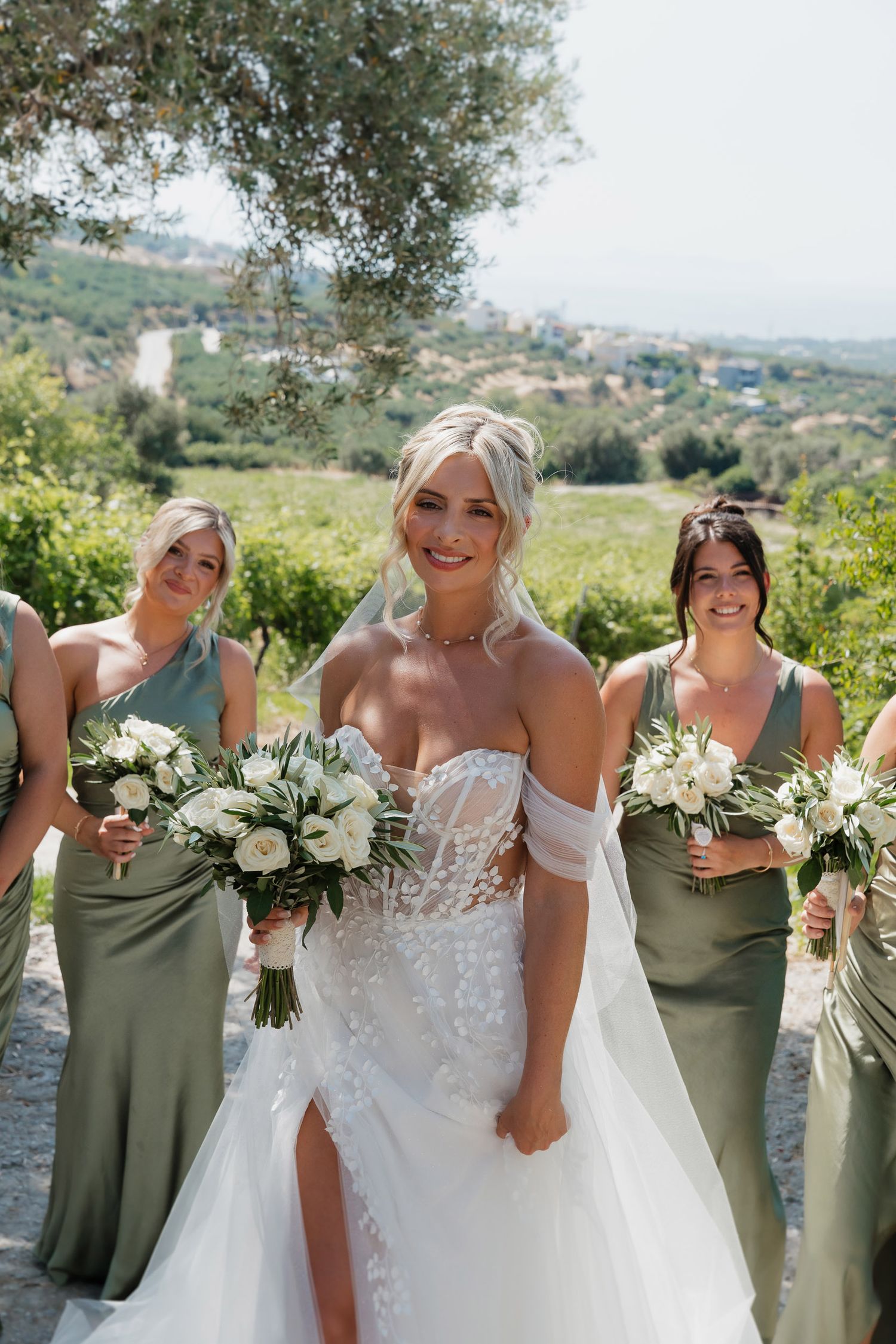 Wedding party members in sage green dresses and white gowns hold bouquets while posing together in a scenic outdoor setting.