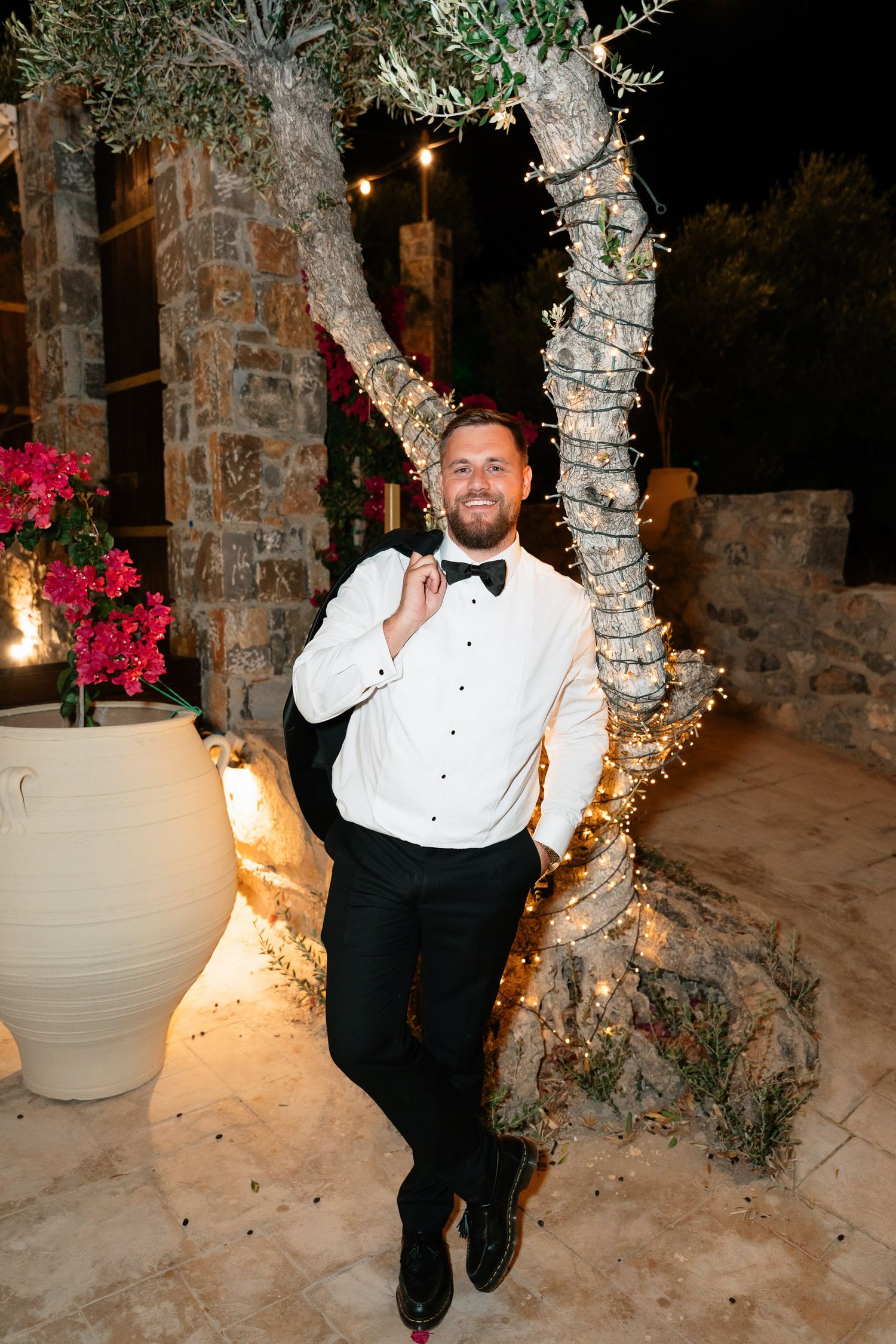A person in formal attire poses playfully in a nighttime garden setting with stone walls and pink flowers.