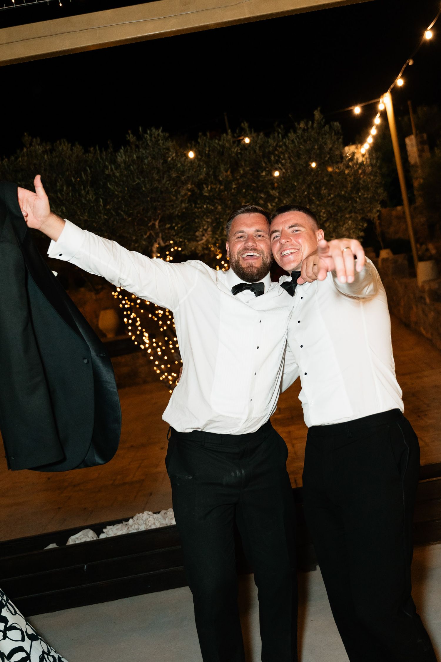 Wedding party members in formal attire dance and celebrate at an outdoor evening reception with string lights.