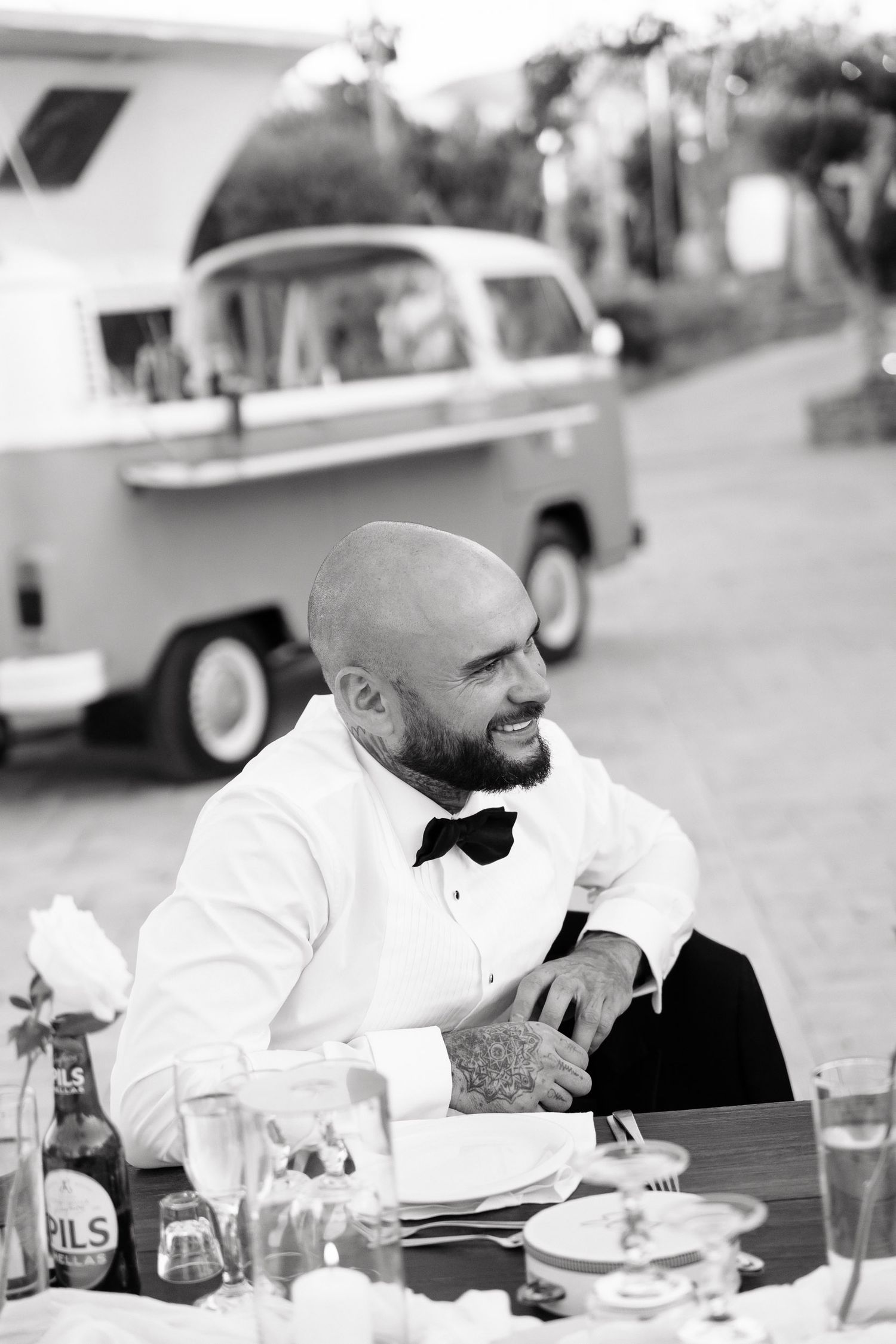 A person in a white dress shirt and bow tie sits at an outdoor dining table with vintage cars visible in the background.