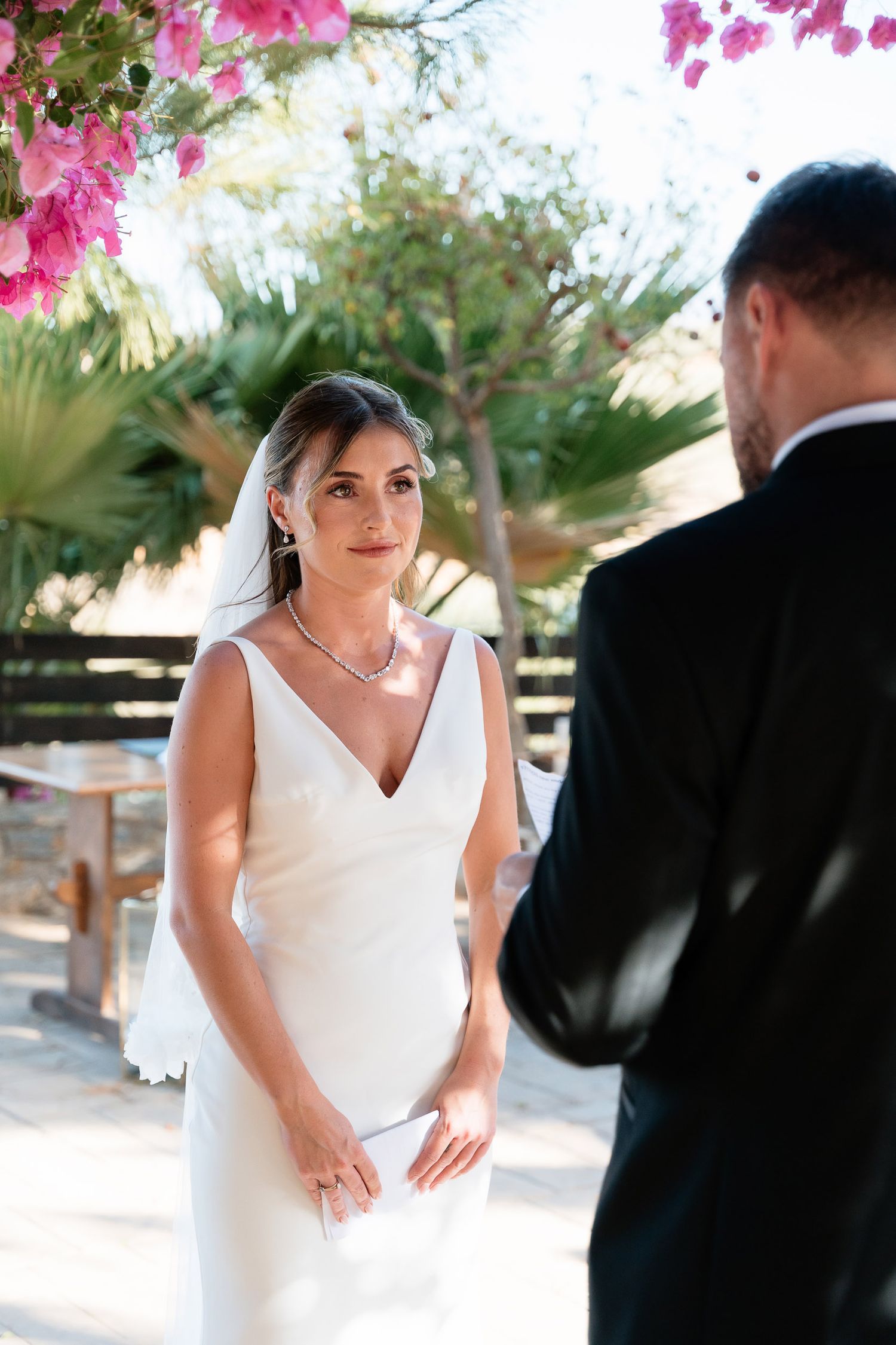 A couple shares a moment in a garden setting with pink bougainvillea flowers overhead and palm trees in the background.