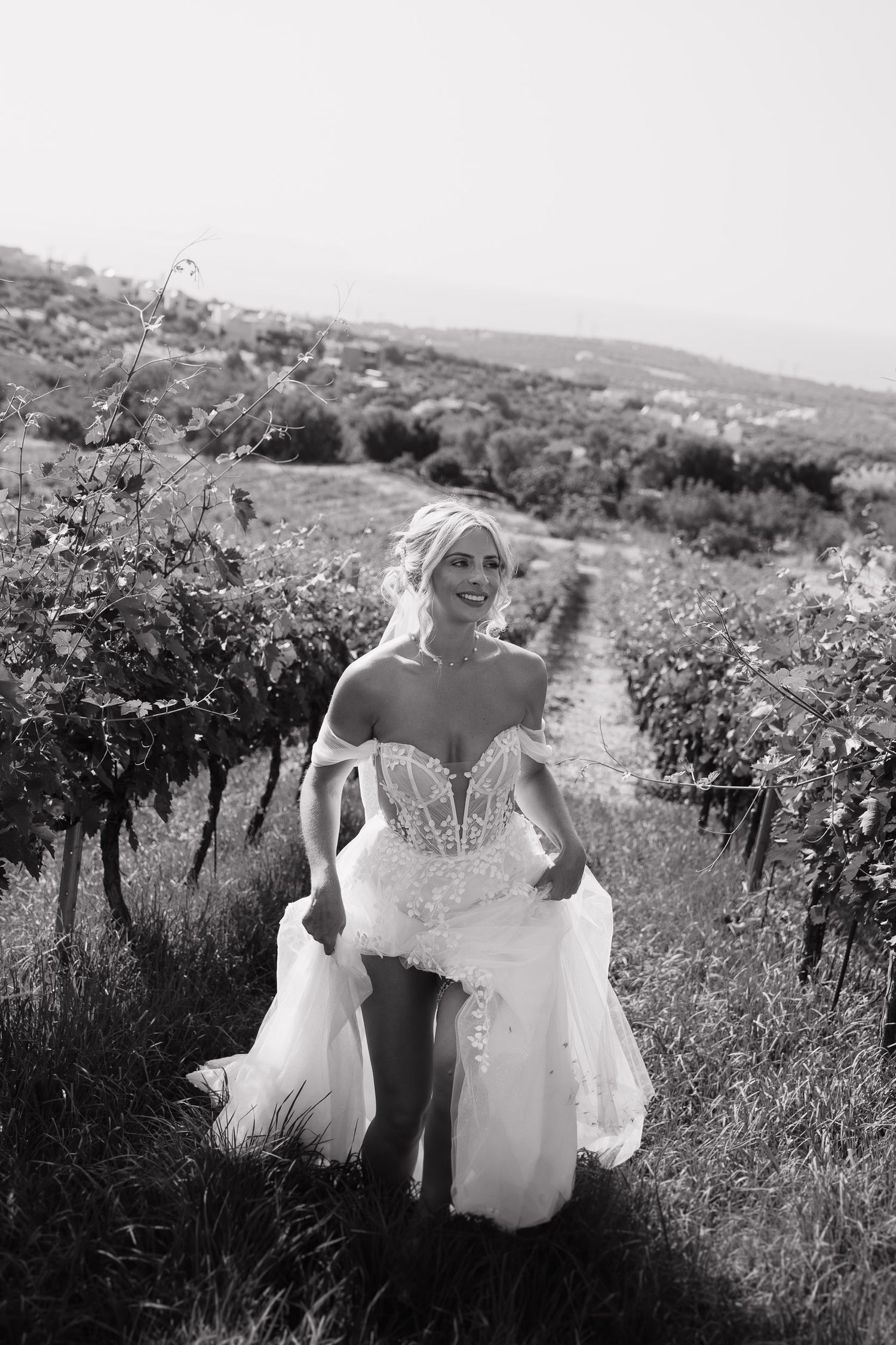 A bride in a white wedding dress walks through a scenic vineyard path on a sunny day.