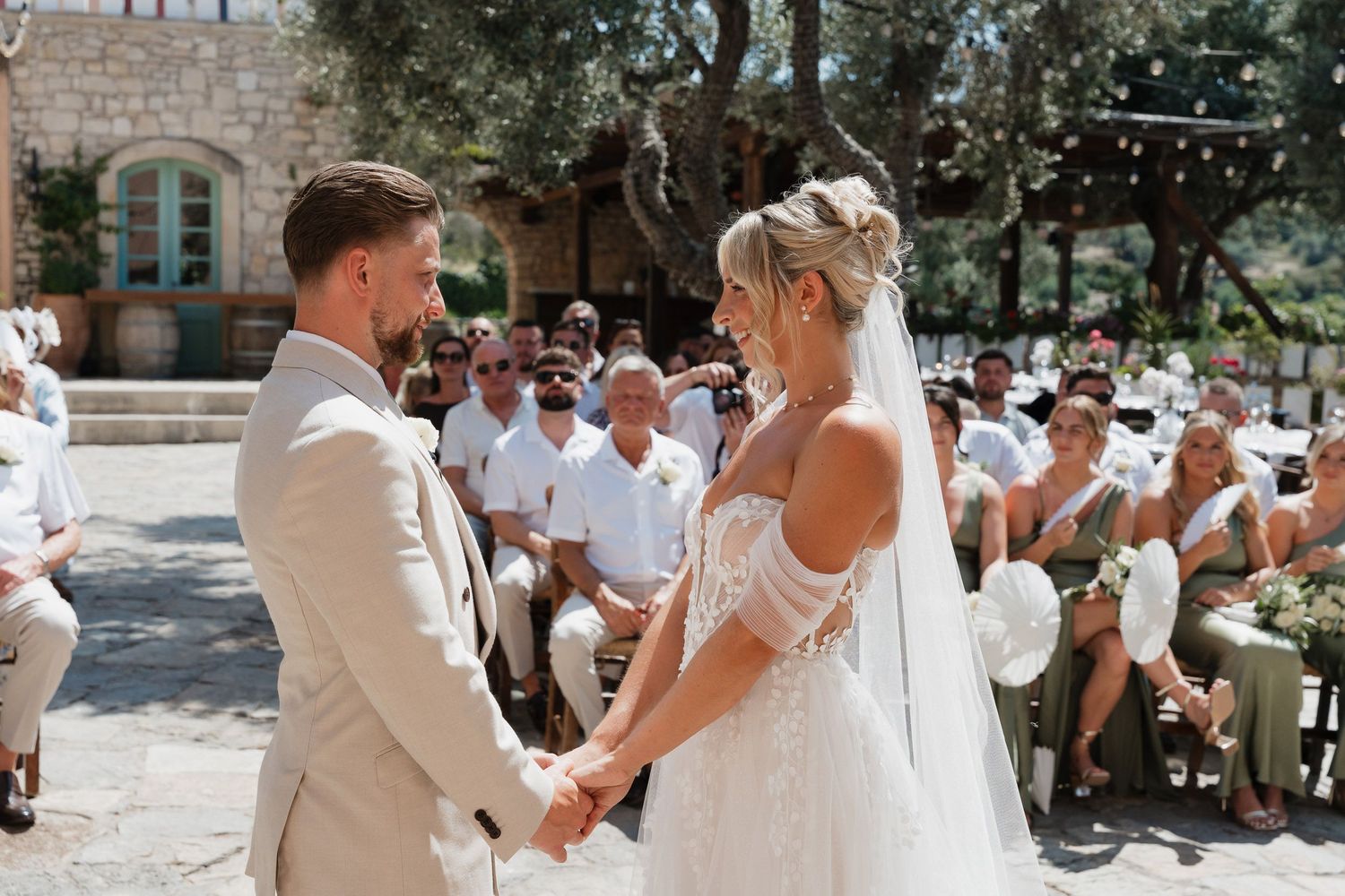 A wedding ceremony takes place outdoors with guests seated in white chairs on a sunny summer day.