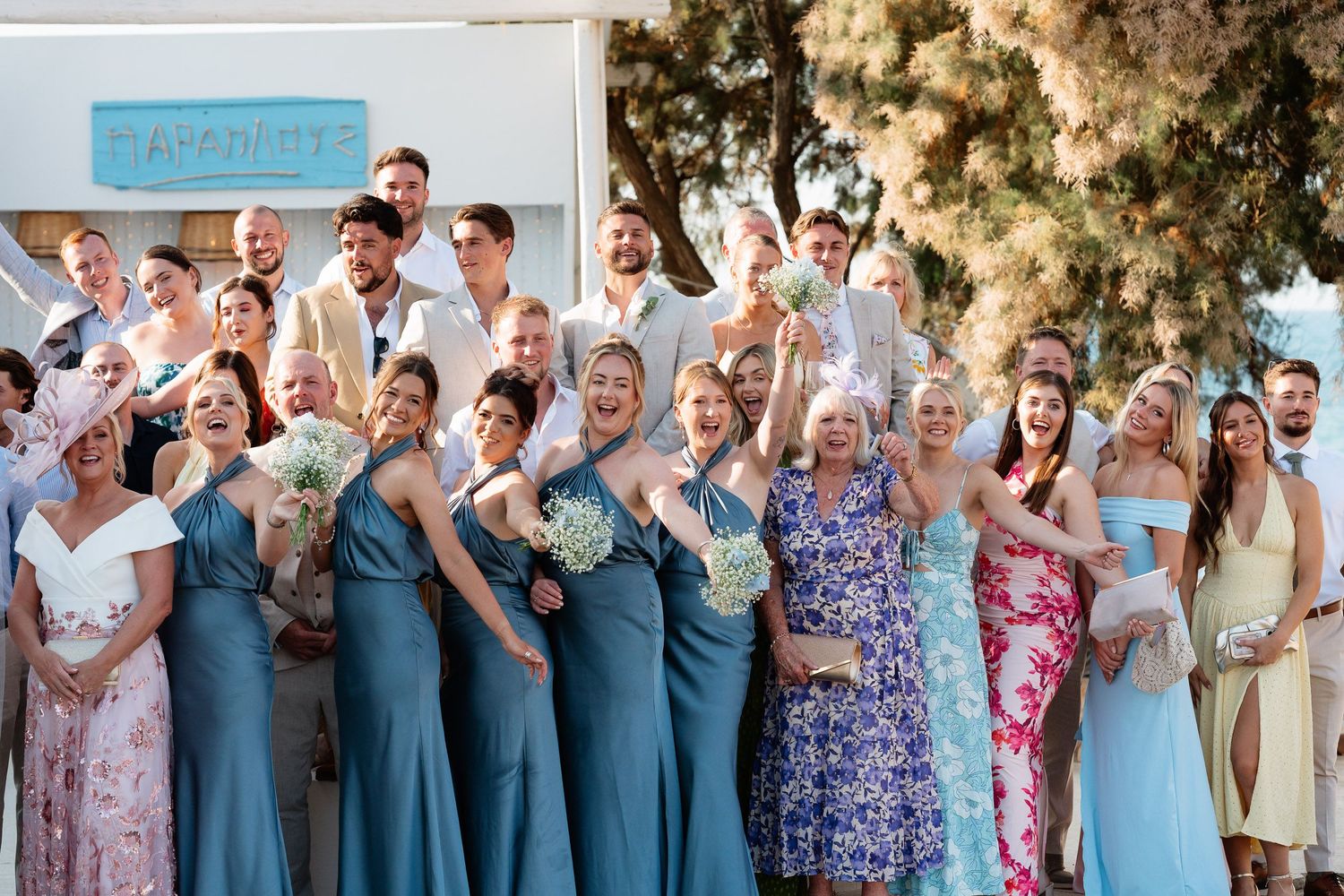 A wedding party celebrates outdoors with joyful poses and coordinated blue and floral dresses.