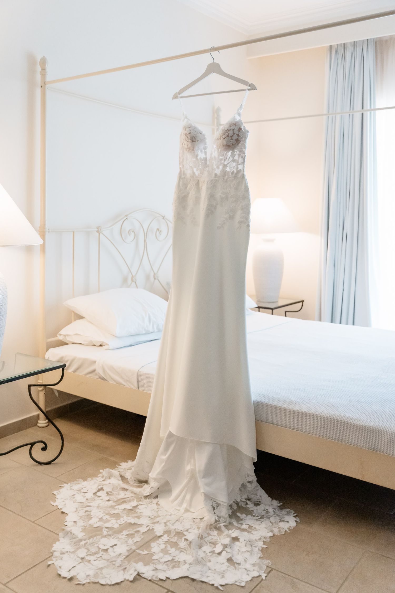 Elegant white lace wedding dress with train hanging on display in a bright bedroom with ornate iron bed frame.