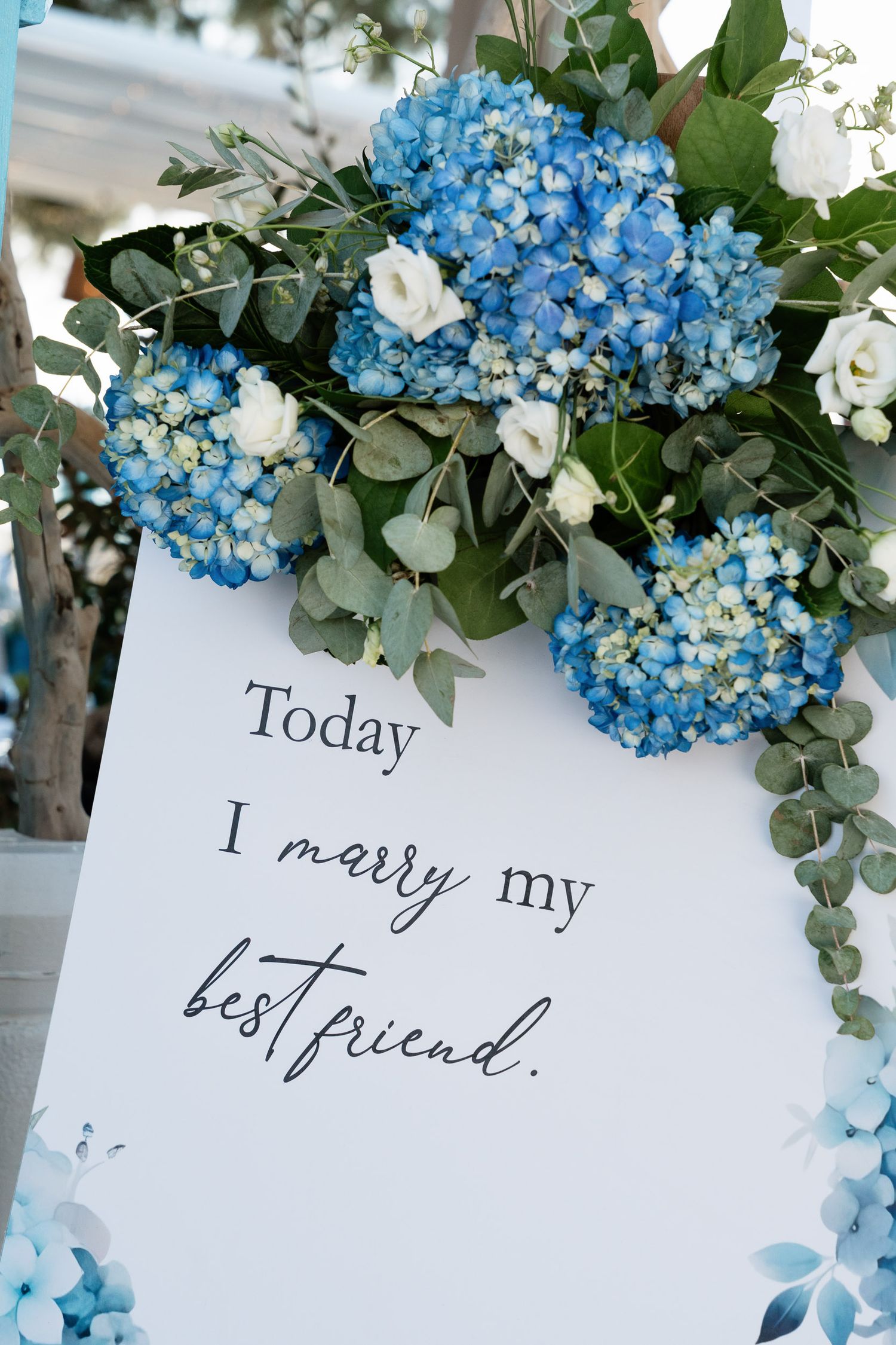 Wedding sign decorated with blue hydrangeas and eucalyptus reads 'Today I marry my best friend' in elegant script.
