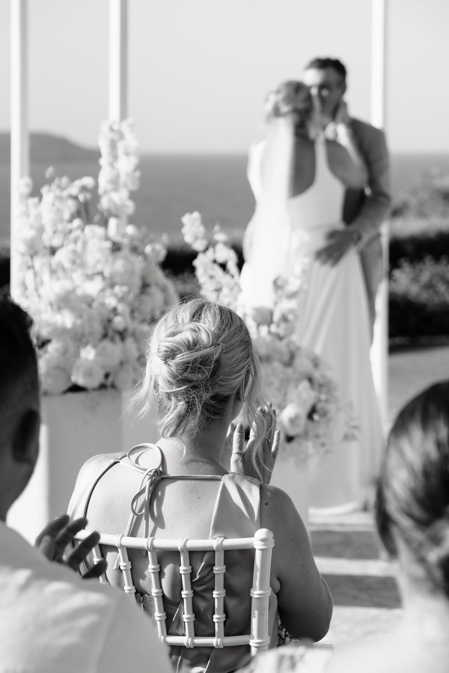 A black and white wedding ceremony photo showing a guest seated in a chiavari chair watching the couple at the altar.