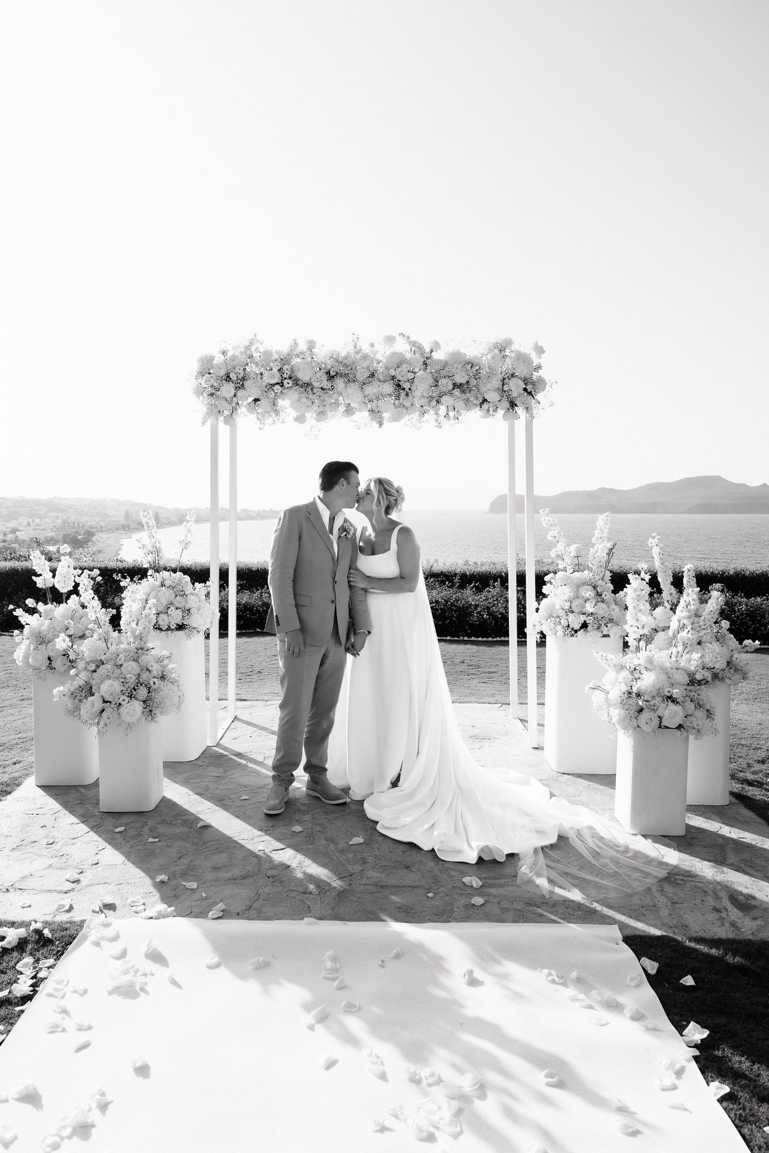 A romantic seaside wedding ceremony with a white arbor and floral arrangements overlooking the ocean at sunset.