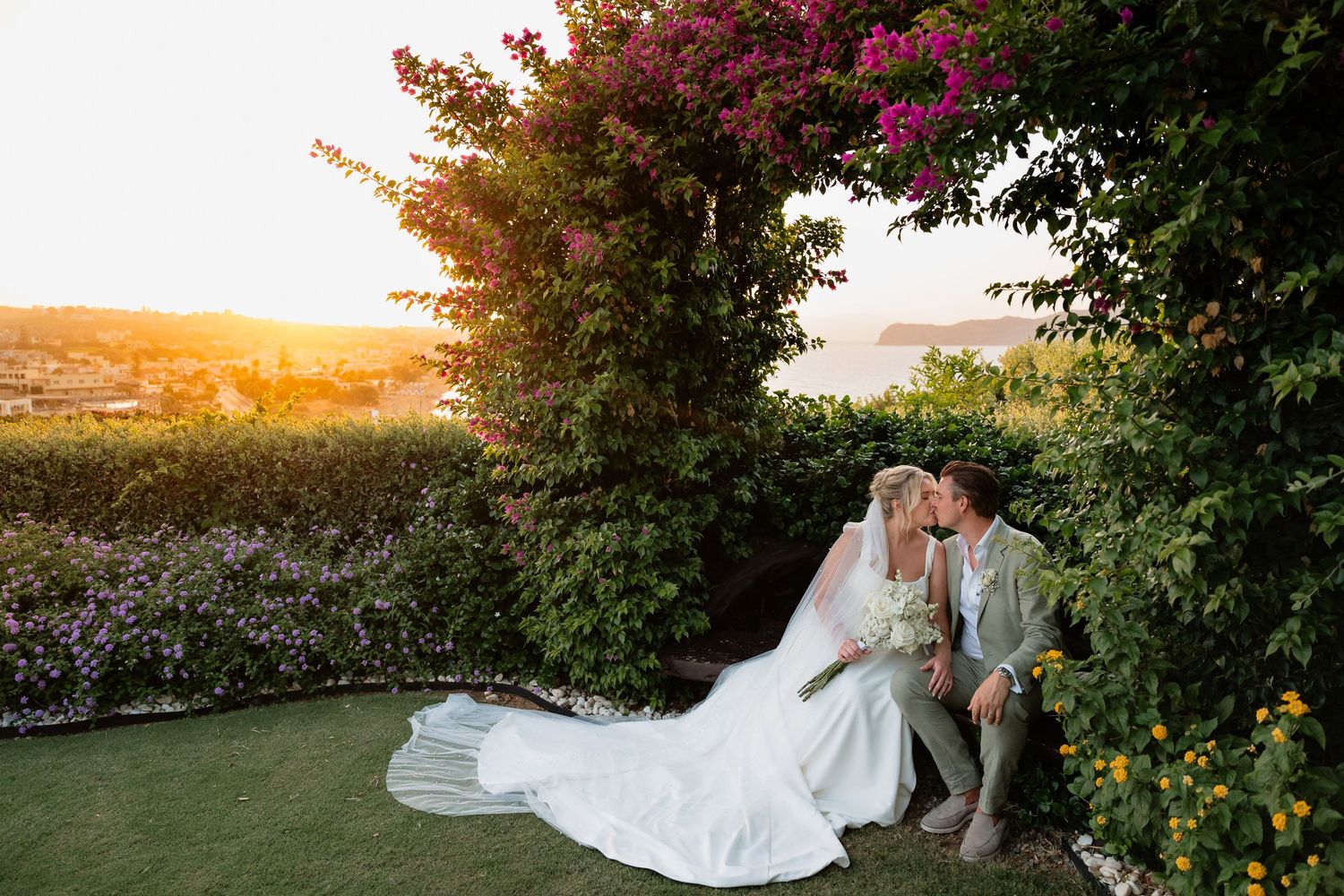 A romantic couple in wedding attire sits together on grass during golden hour sunset with flowering bushes in background.