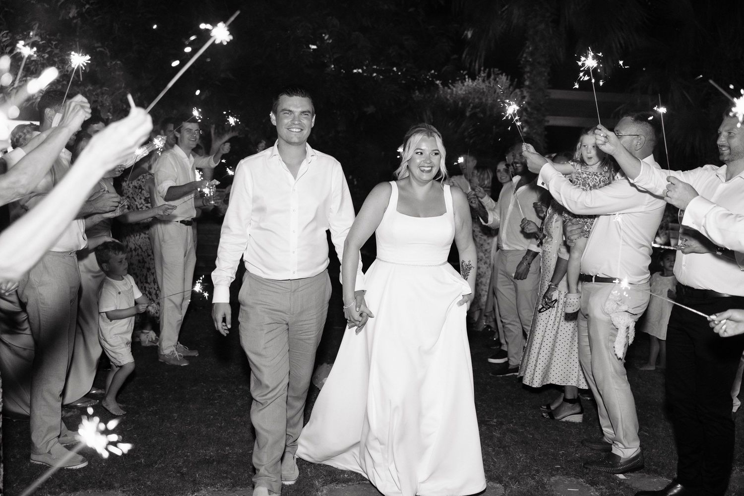 Wedding couple exits their reception through a sparkler tunnel lined with guests in black and white photography.