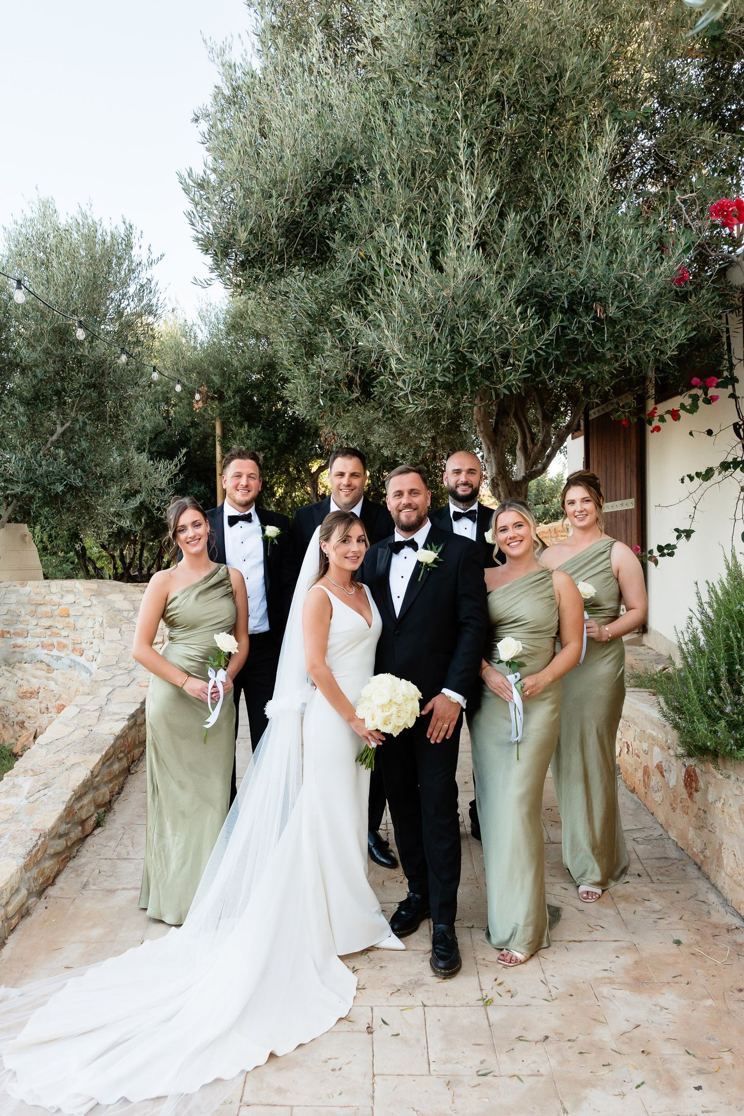 Wedding party poses playfully against stone wall with olive trees in Mediterranean garden setting.