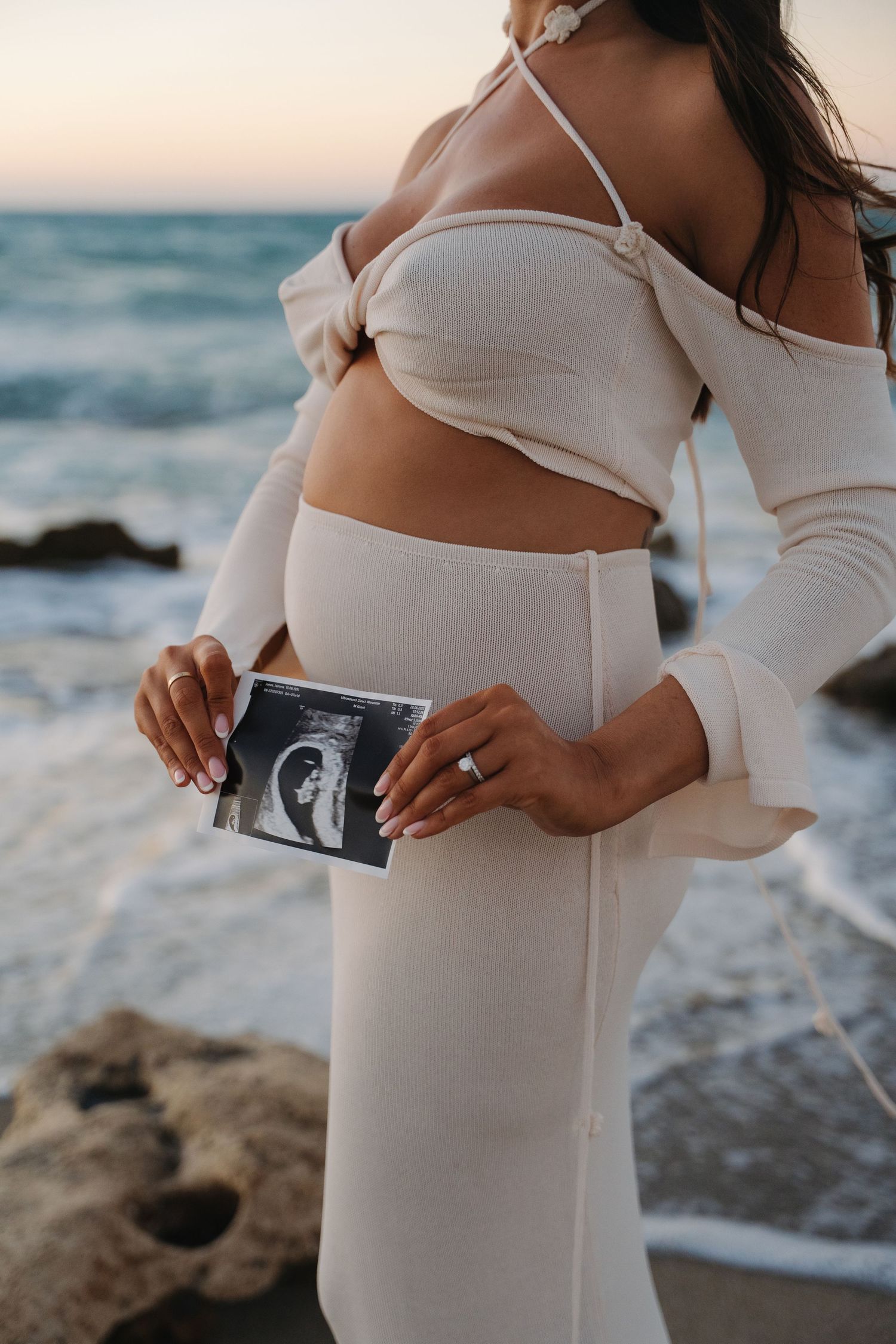 A person in a white dress holds ultrasound photos while standing on a rocky beach at sunset.