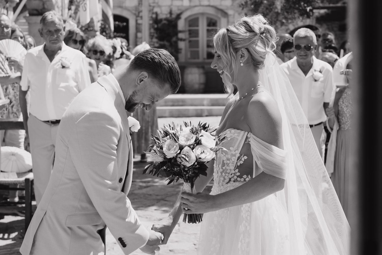 A black and white photograph captures a romantic moment between a couple in elegant white attire at an outdoor wedding ceremony.